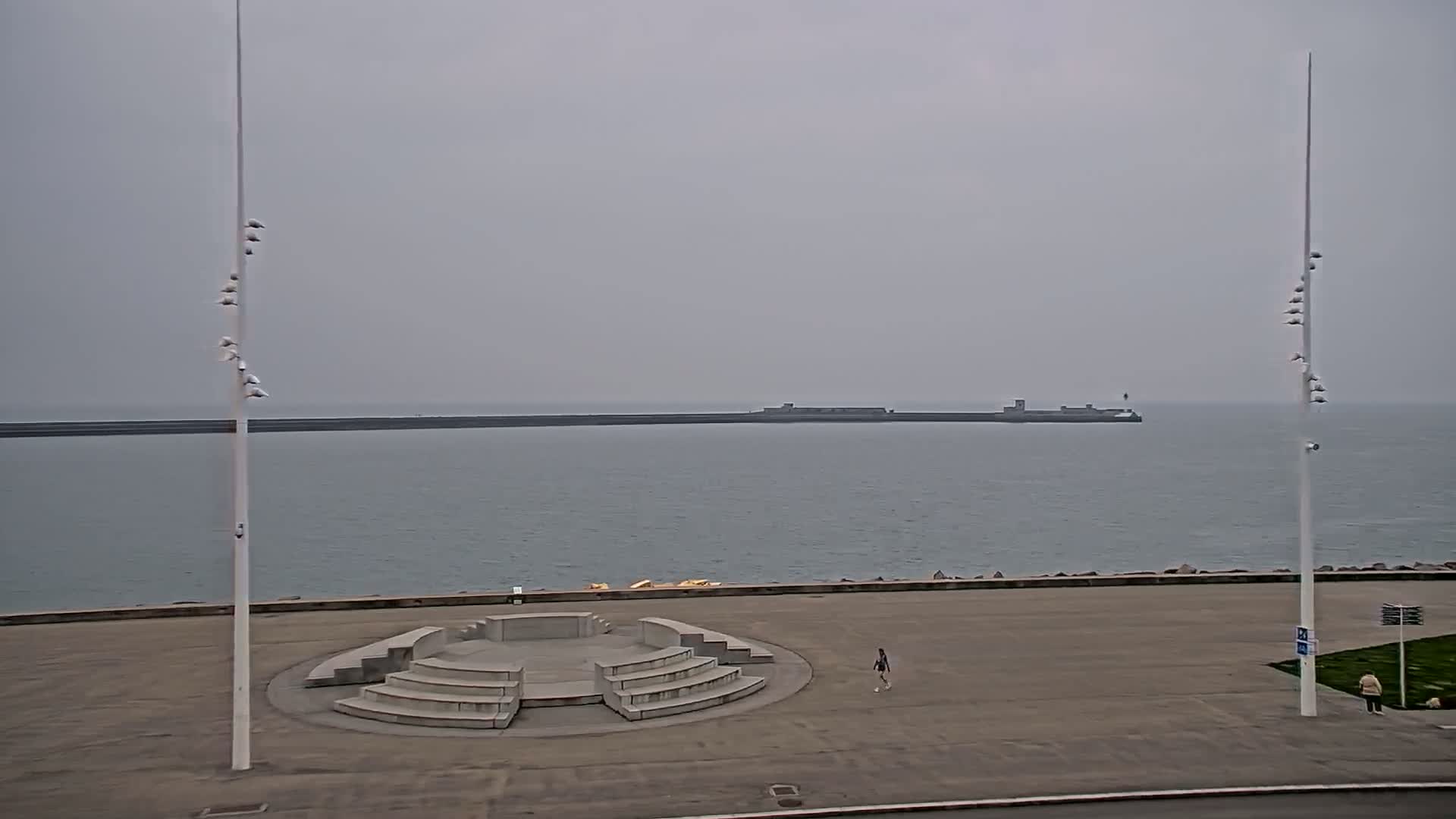 A gray, overcast day shows a calm body of water with a distant breakwater, a paved area with a stone amphitheater structure and two tall light poles, and two lone pedestrians.