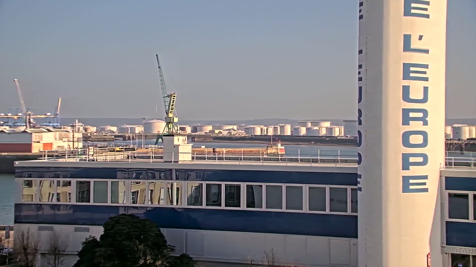 A clear, sunny day reveals a harbor scene with industrial buildings, storage tanks, cranes, and a large structure with a blue facade reflecting the surroundings.
