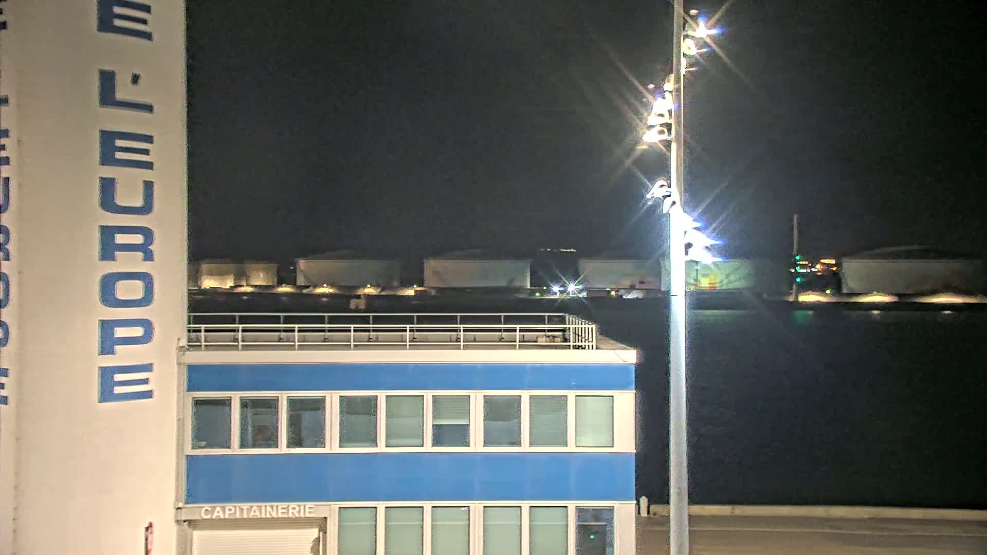 A nighttime view shows a blue building with a rooftop railing, a tall white structure with blue lettering, a bright light post, and several large, light-colored structures in the background across dark water.