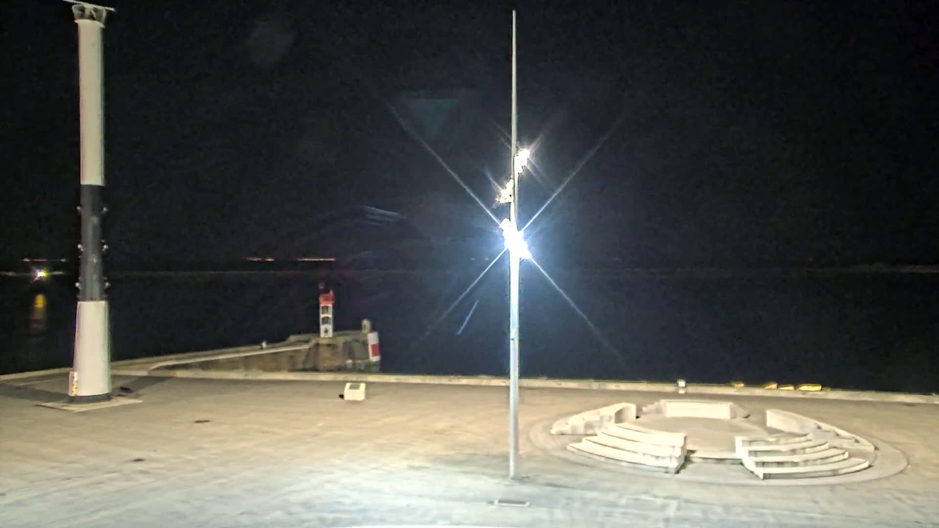 A nighttime view of a harbor, featuring a tall, striped lighthouse, a smaller lighthouse in the distance, a circular amphitheater-like structure, and a bright lamppost.