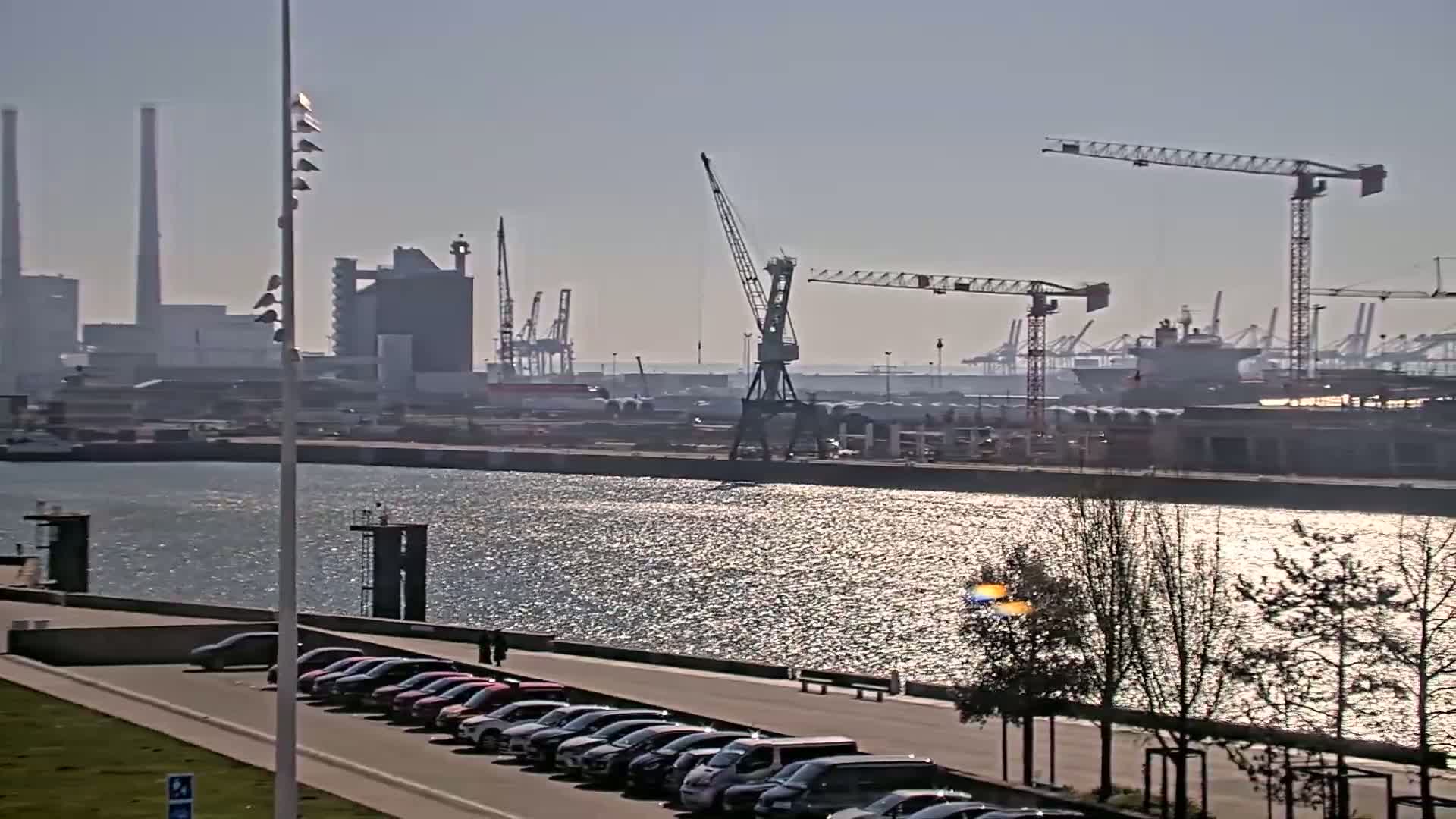 A sunny day at a harbor shows several cranes, industrial buildings, and ships across a body of water, with parked cars visible along a waterfront walkway.
