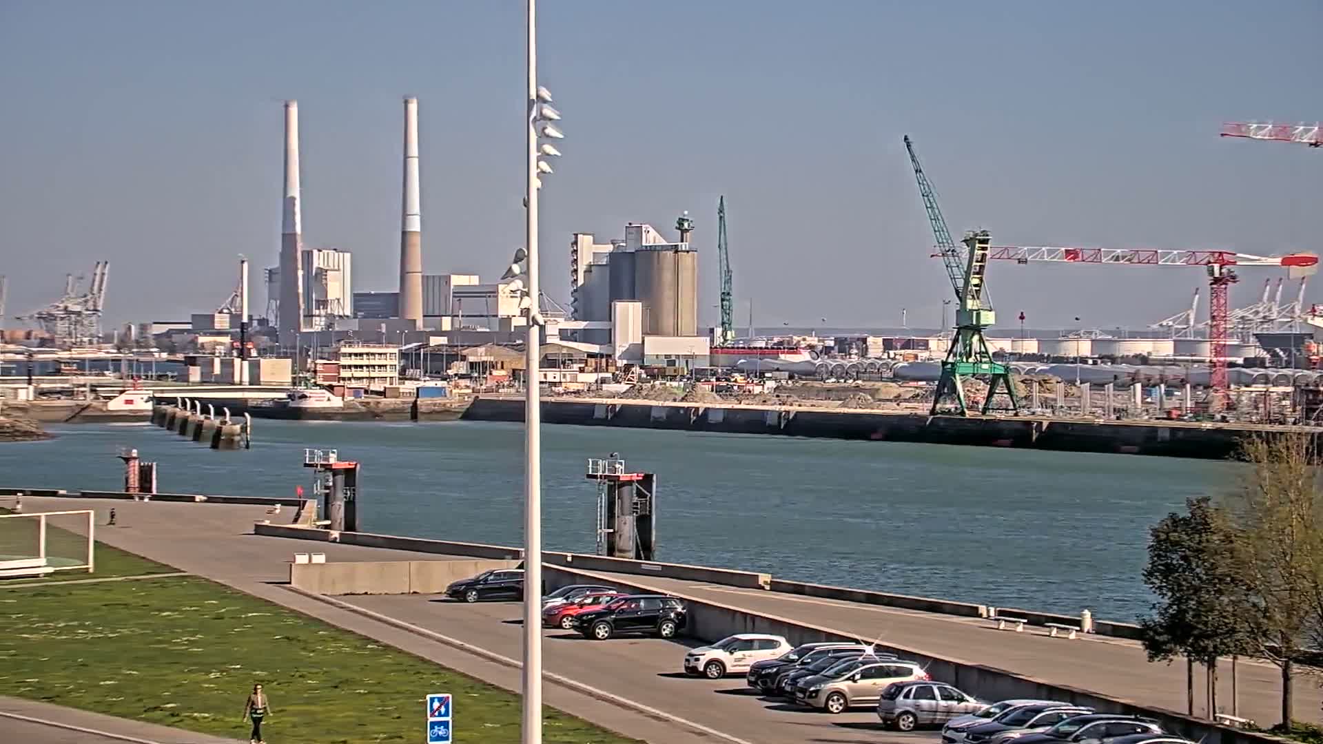 A sunny day reveals a harbor scene with industrial buildings, cranes, and parked cars along a paved waterfront path next to a calm body of water.