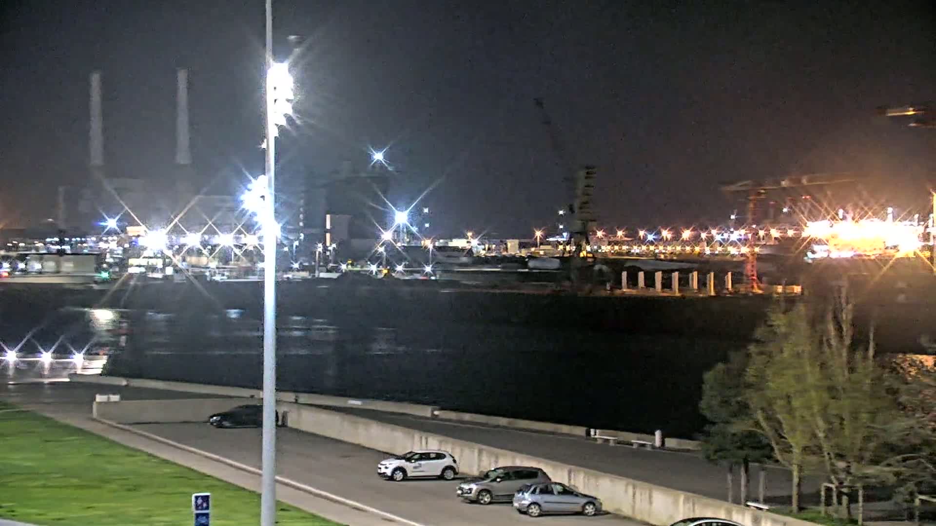 At night, a brightly lit industrial port area is seen across a body of dark water from a paved walkway with several parked cars and small trees.