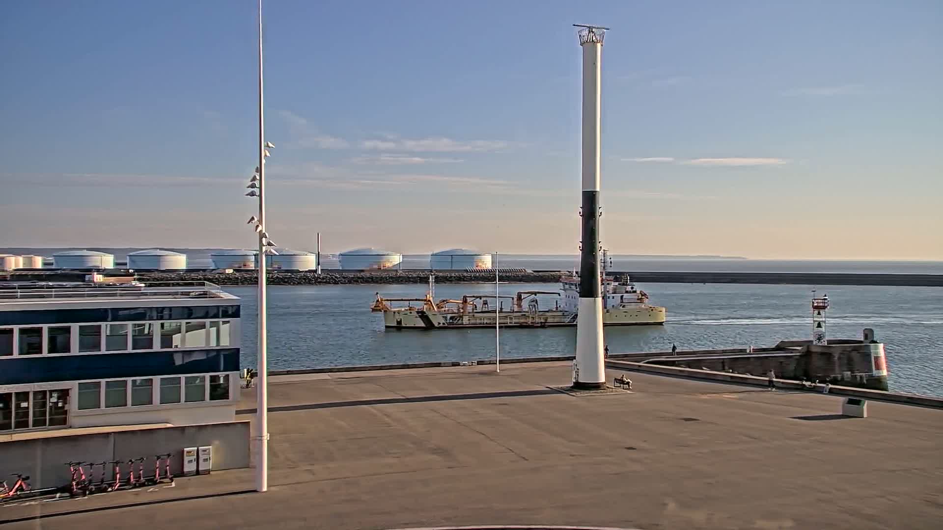 A harbor scene on a clear, sunny day shows a dredging vessel in the water near a tall, slender lighthouse, with storage tanks and a building visible on the shore.