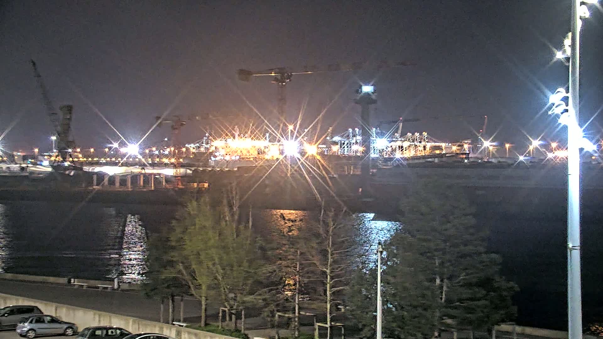 A brightly lit nighttime industrial port scene with cranes and ships is viewed from across a body of water, with several small trees in the foreground.