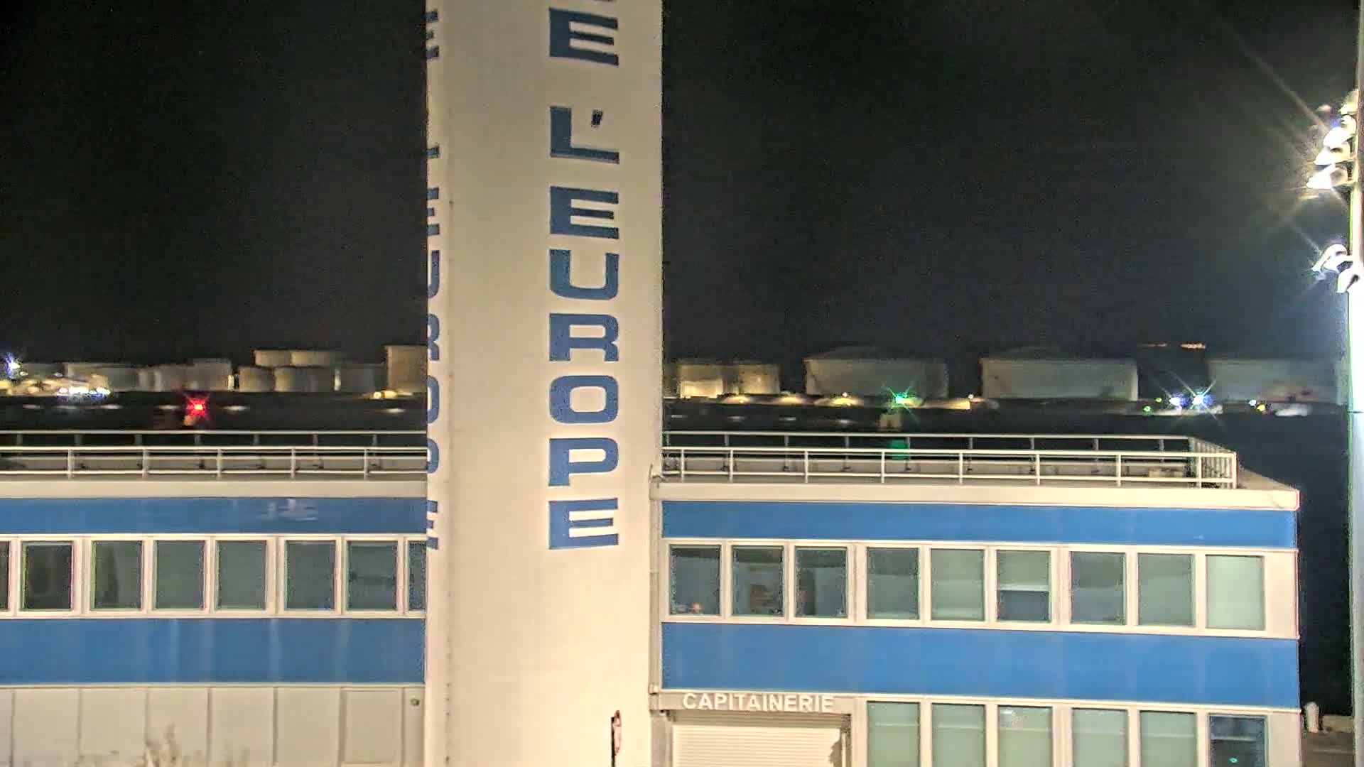 At night, a tall, light-colored structure with "L'EUROPE" vertically written on it stands next to a blue building with many windows, behind which are several industrial storage tanks.