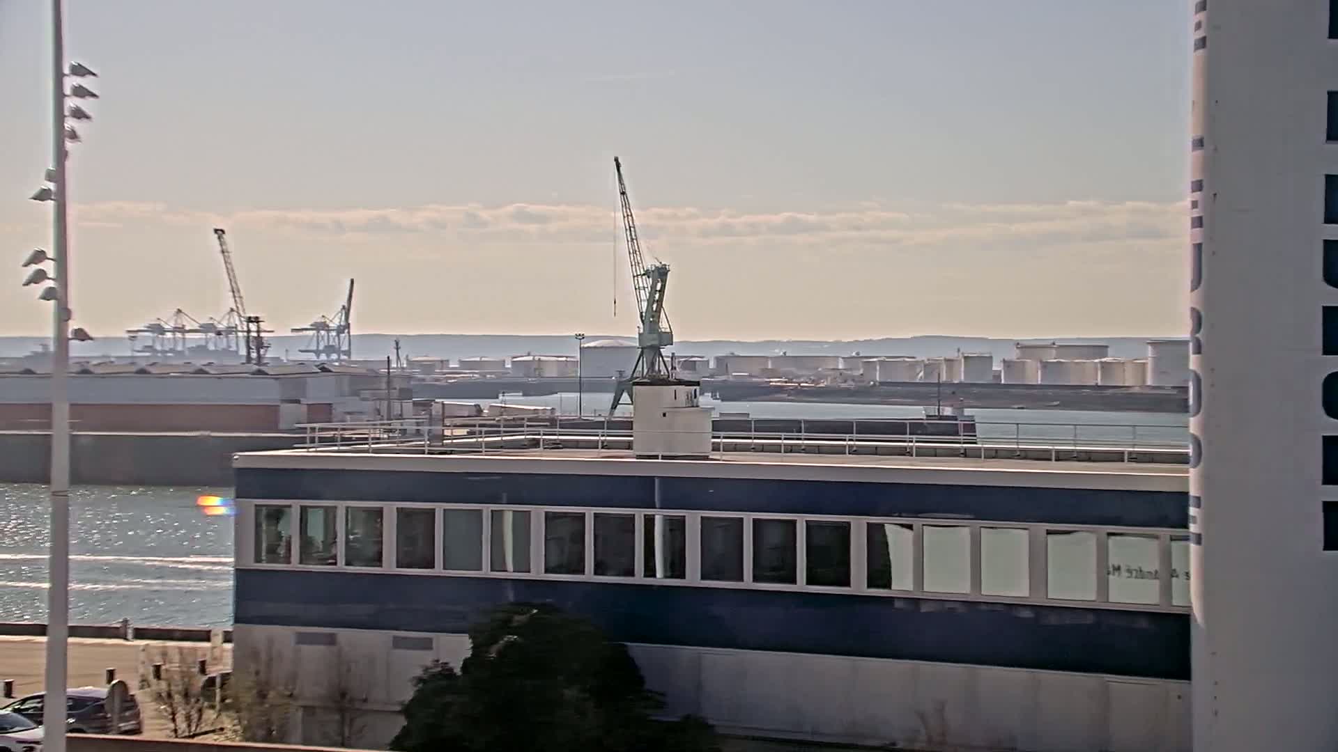 A blue building with many windows sits on a waterfront overlooking a harbor with cranes, industrial storage tanks, and calm water under a mostly clear sky.