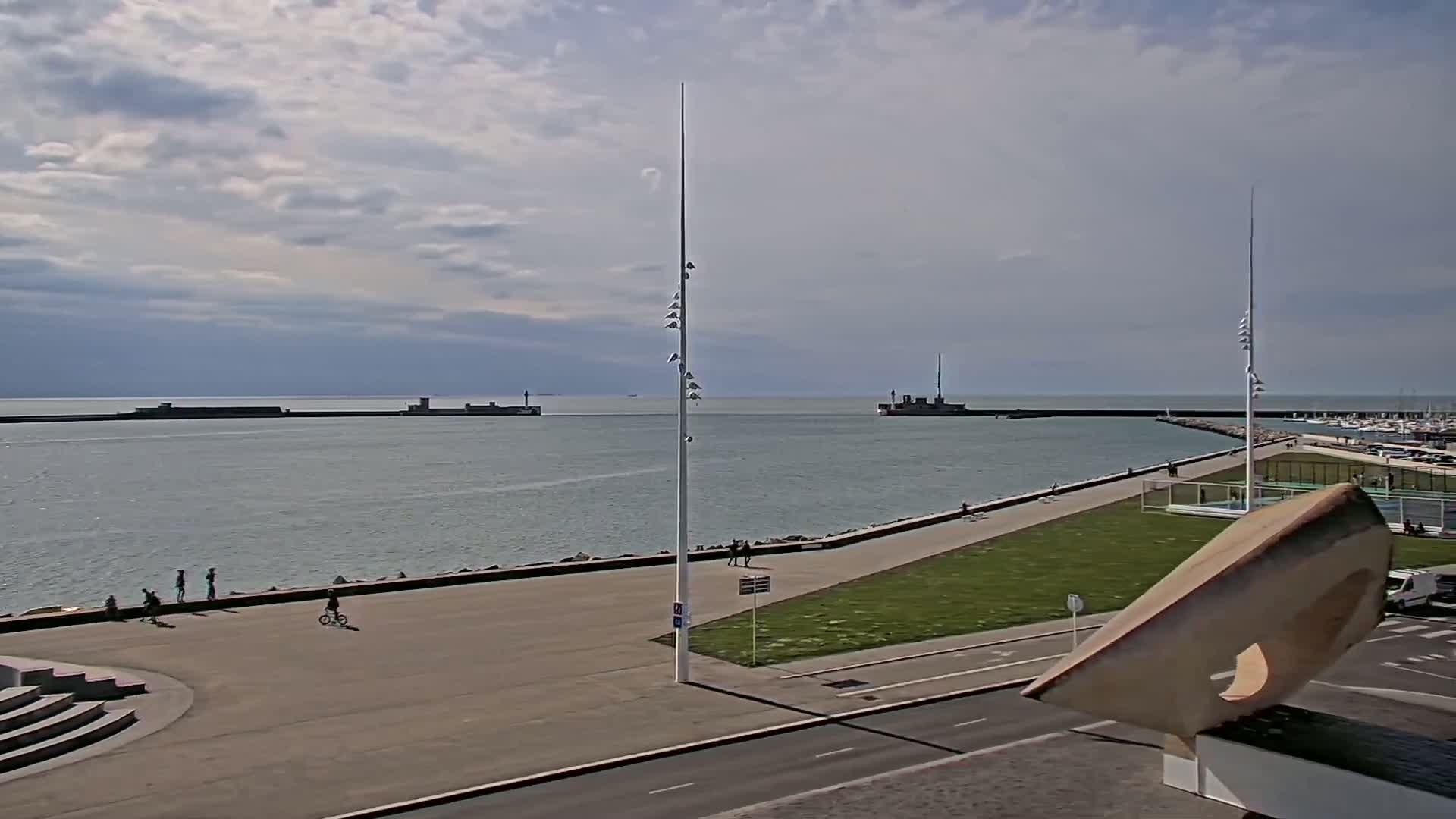 A partly cloudy day reveals a harbor with calm water, a paved walkway lined with people, and a large, abstract sculpture in the foreground.