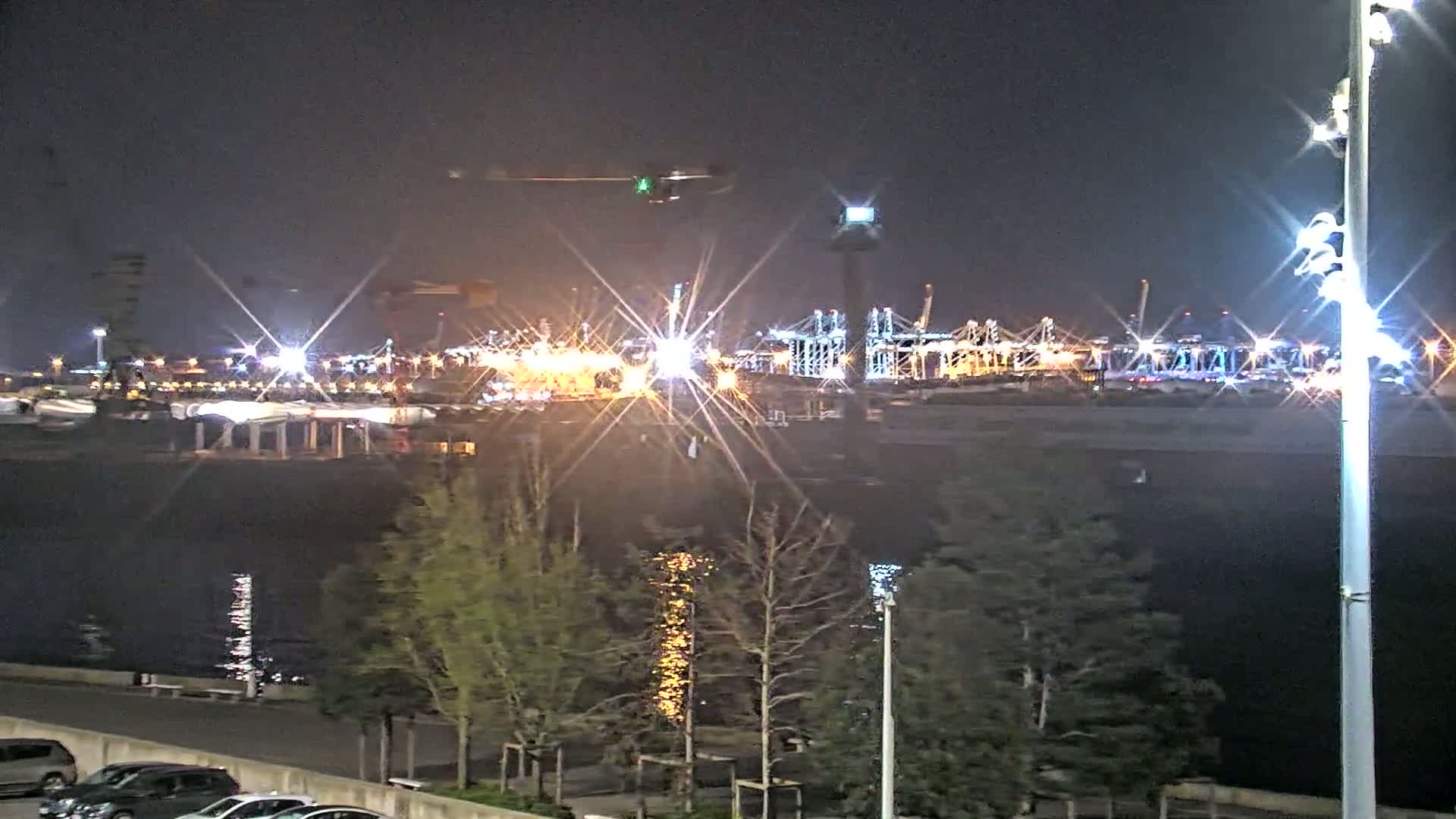 A brightly lit nighttime view of a harbor filled with cargo ships and cranes, seen across a body of dark water from a park-like area with small trees and parked cars.