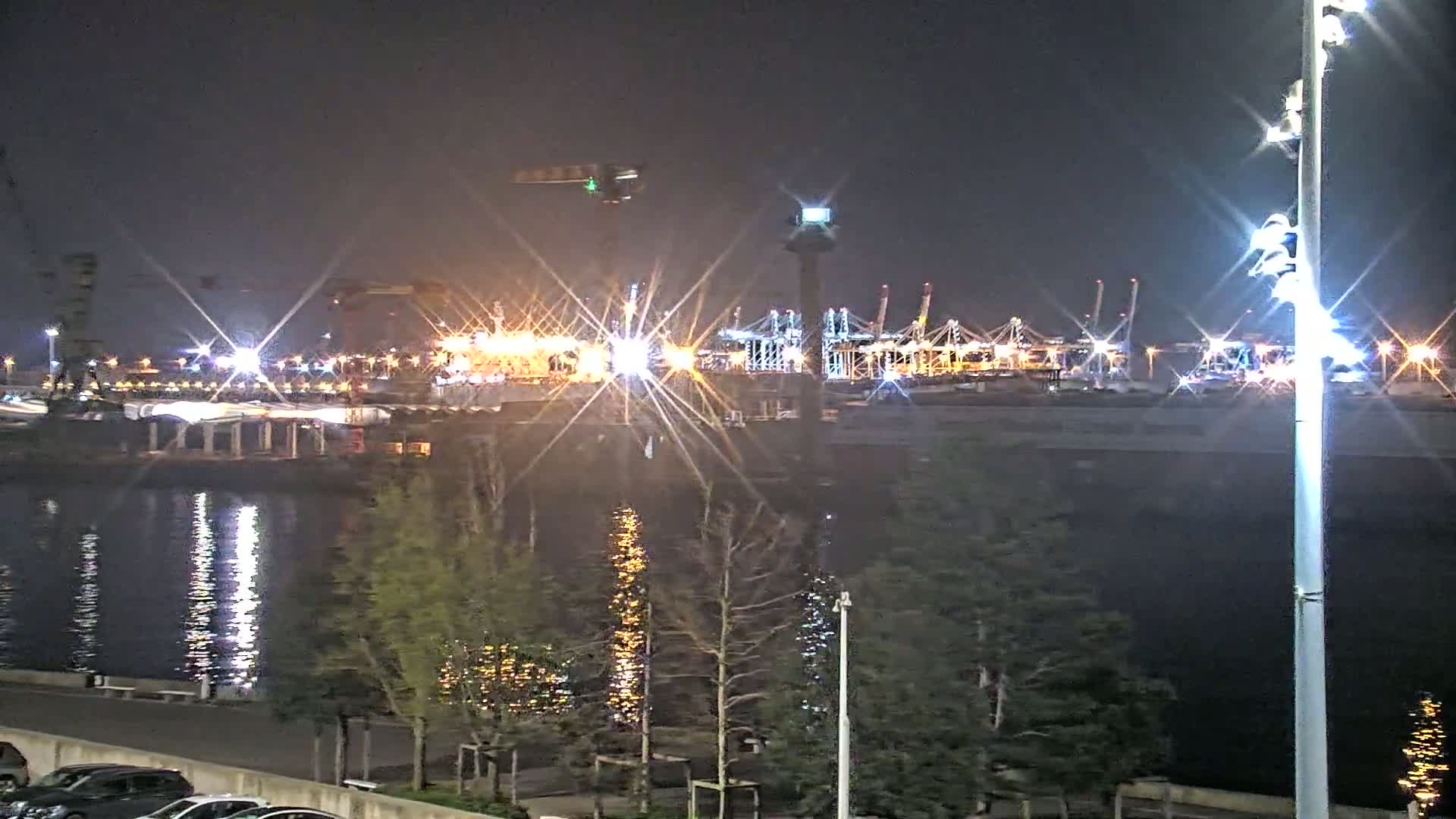 A brightly lit nighttime port with numerous cranes and ships is visible across a body of water from a vantage point lined with trees and parked cars.