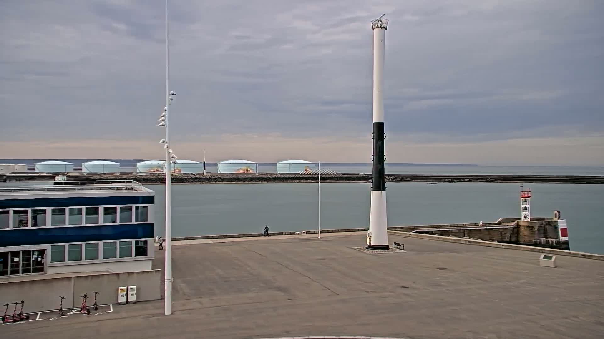 A tall, black and white lighthouse stands in a harbor next to a smaller lighthouse, under an overcast sky, with several large storage tanks visible in the background.
