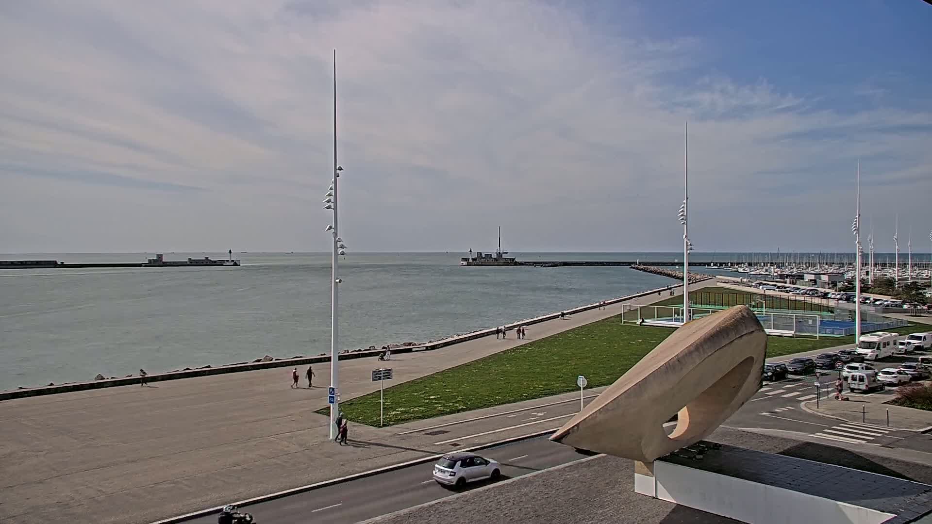 A partly sunny day overlooks a harbor with a marina full of boats, a paved walkway with people strolling, and a grassy area with a unique stone sculpture.