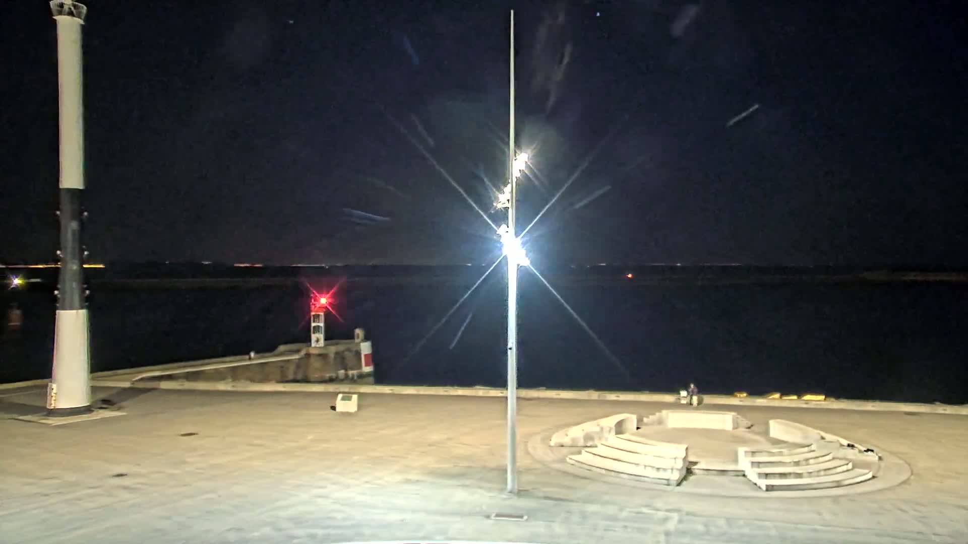 A nighttime view of a harbor with a tall lighthouse, a smaller red light, a tall light post, and a circular stone seating area.