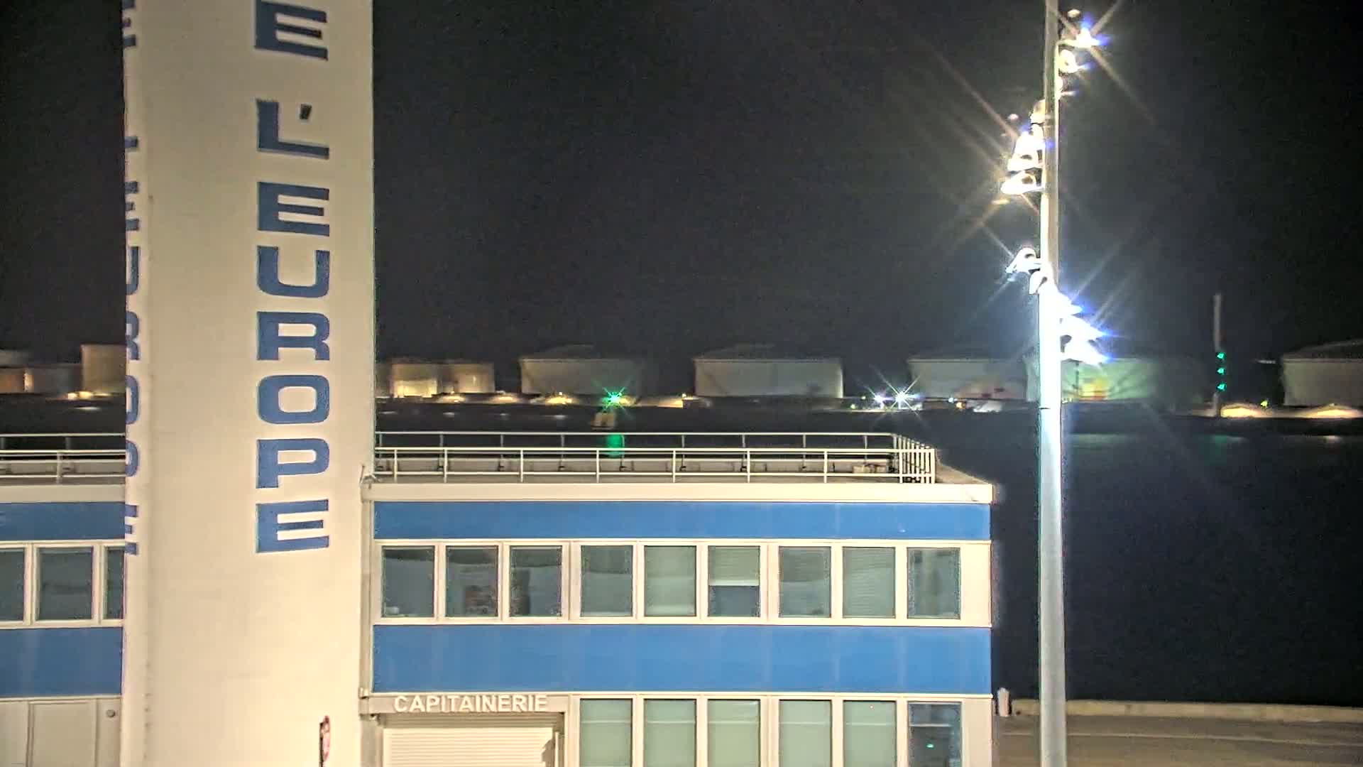 A tall white tower with blue lettering, a blue building with many windows, and bright lights illuminate a nighttime scene.