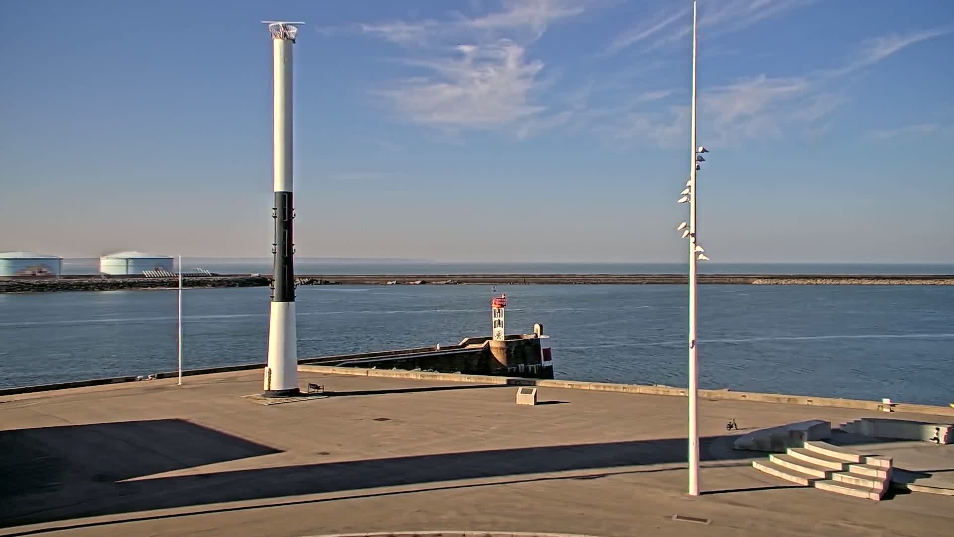 A mostly sunny day shows a harbor with a tall white and black striped tower, a smaller lighthouse, and a flagpole, all situated on a paved area next to calm blue water.