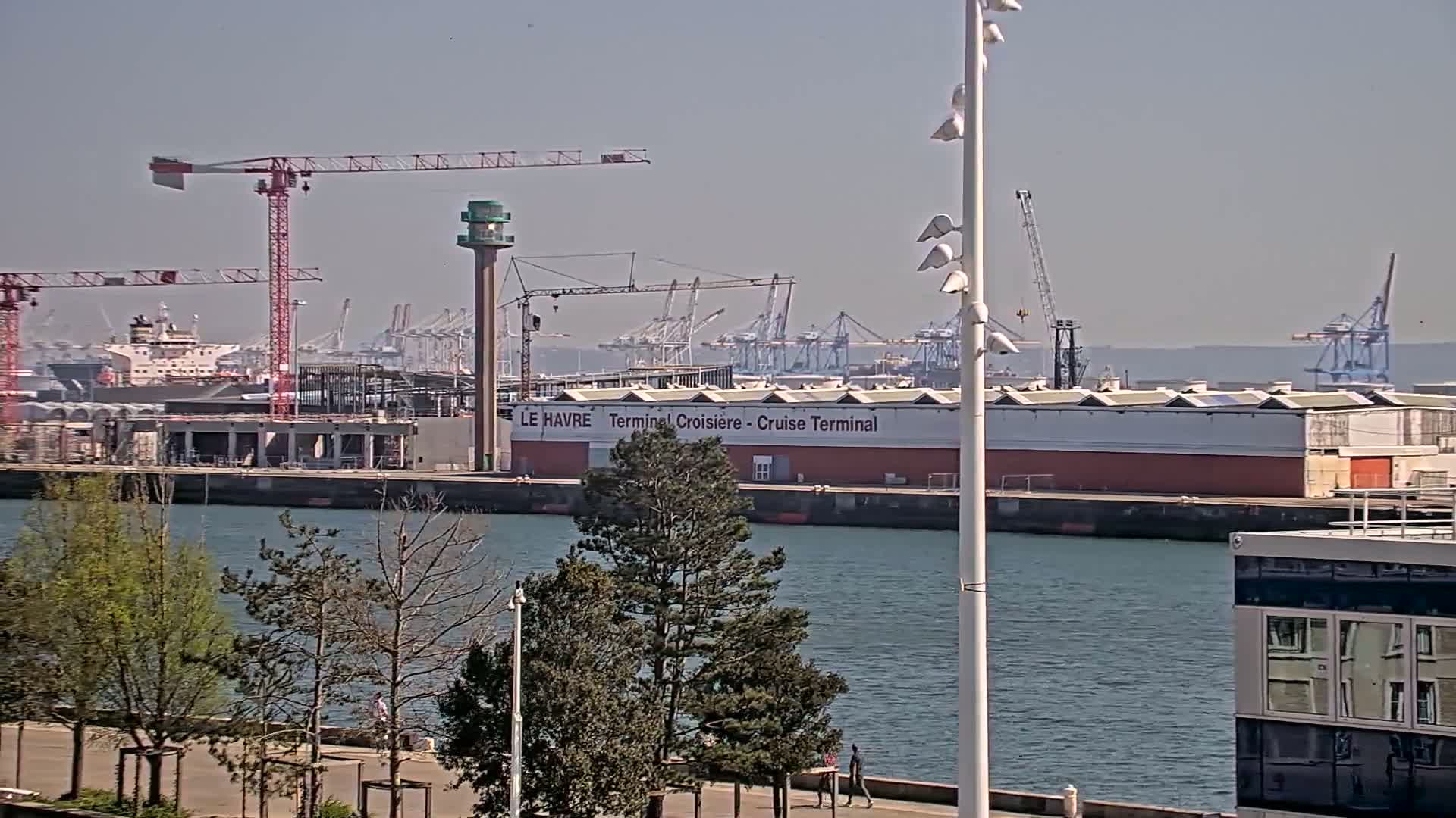 A harbor scene on a clear, sunny day shows a cruise terminal, numerous cranes, ships, and a calm body of water with trees and a walkway in the foreground.