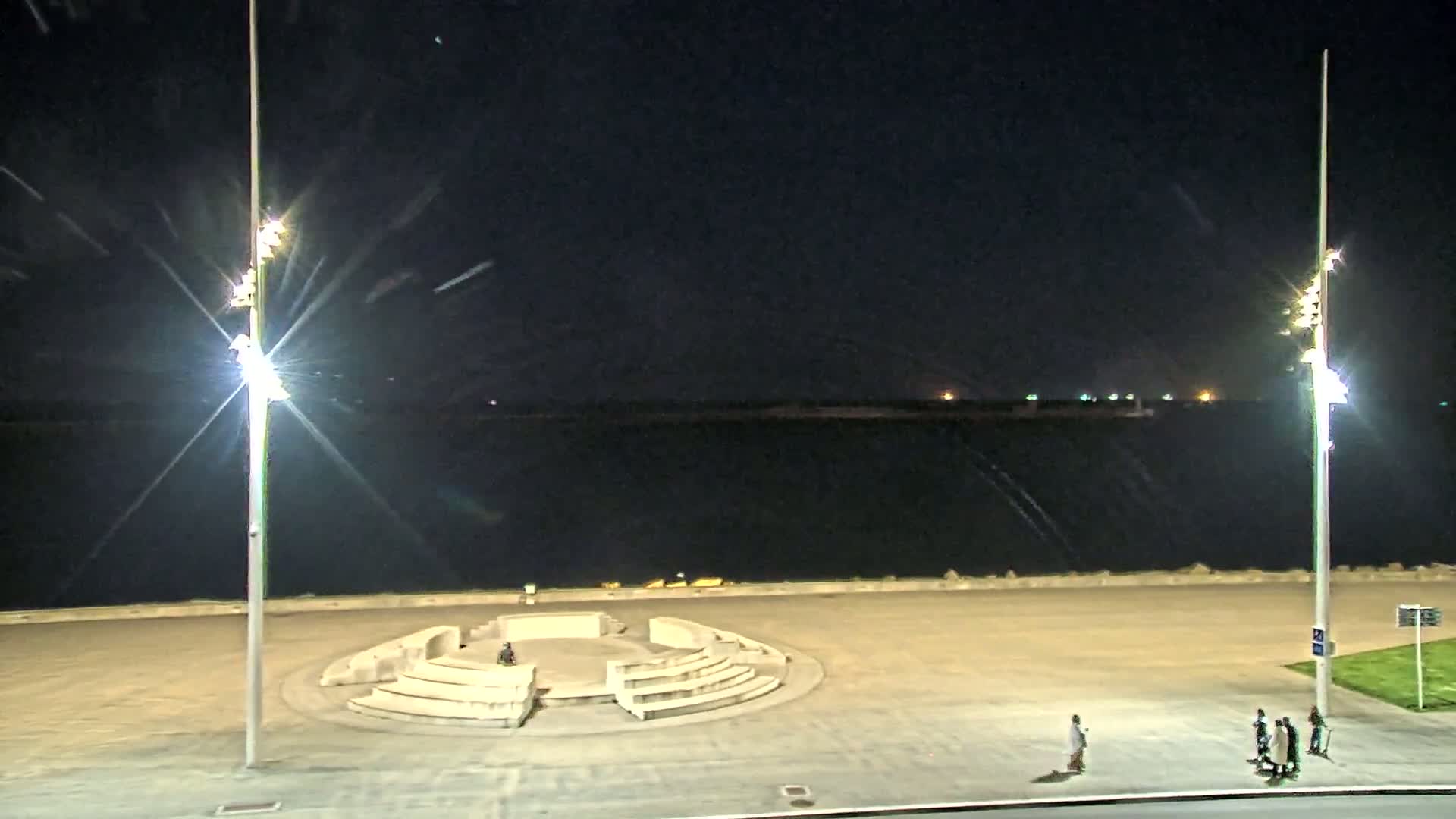 At night, a circular stone amphitheater sits on a paved waterfront area overlooking a dark body of water under bright lights.