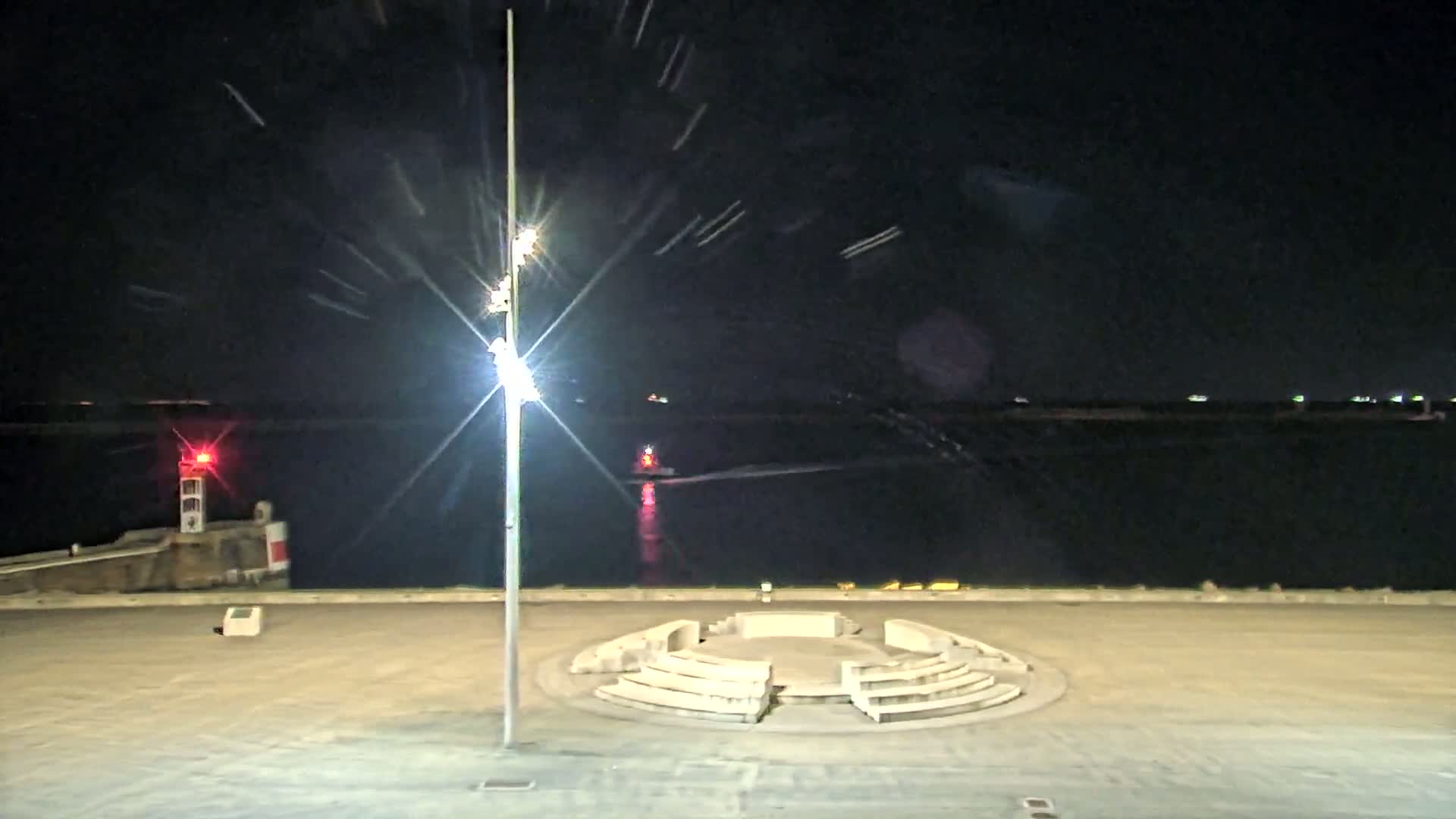 A brightly lit nighttime scene shows a circular stone seating area overlooking a calm body of water with a small boat and a red light in the distance.