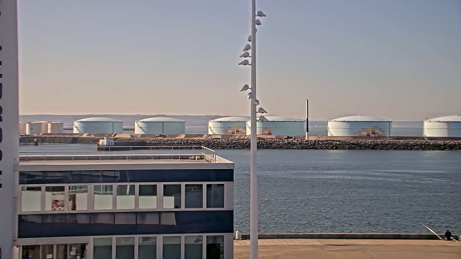 A row of large, light-blue storage tanks sits across a body of calm water from a dark-blue building, under a clear sky.