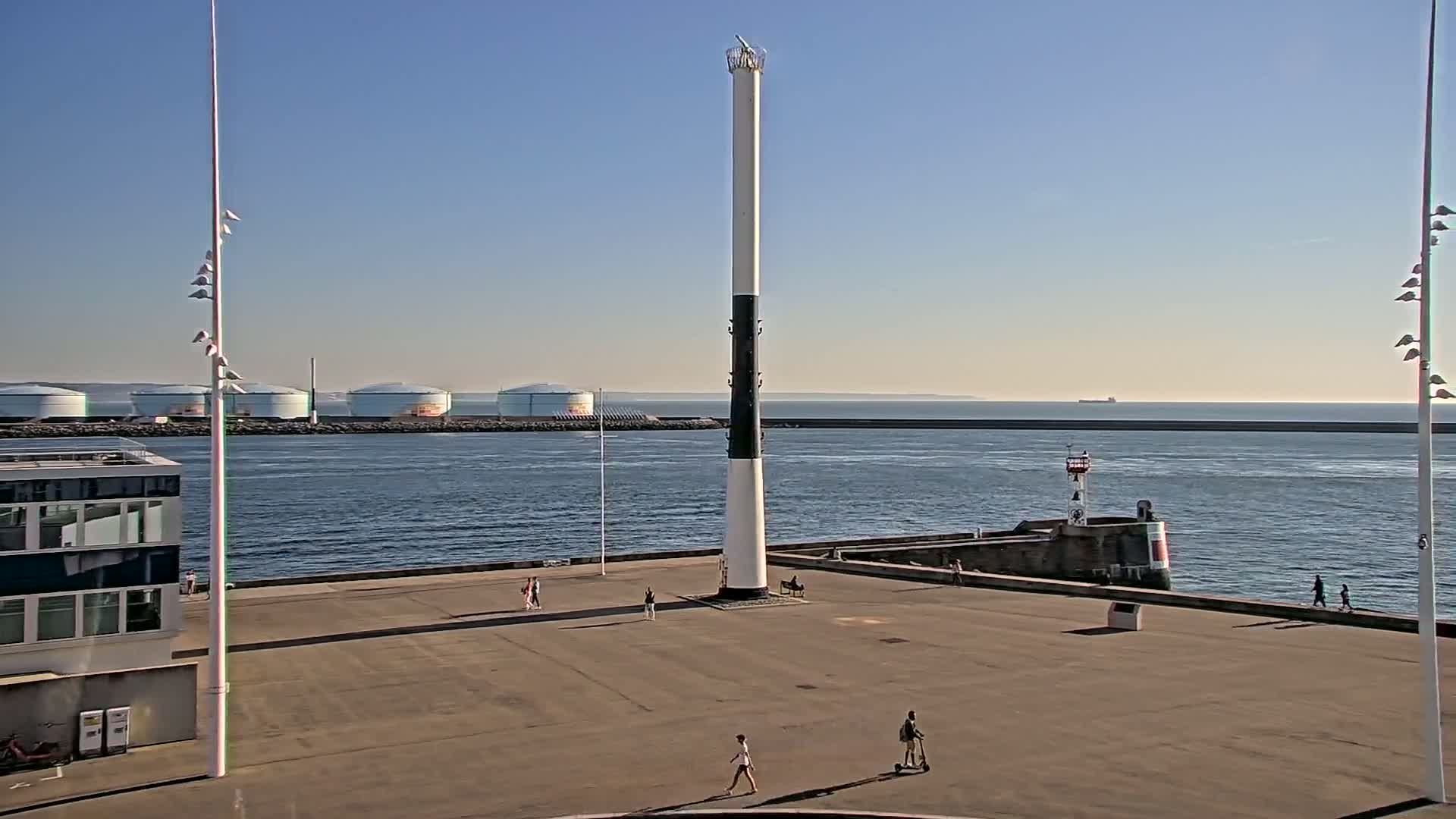 A tall, black-and-white lighthouse-like structure stands in a paved area overlooking calm water, with a few people strolling nearby, under a clear, sunny sky; several large storage tanks are visible in the distance.
