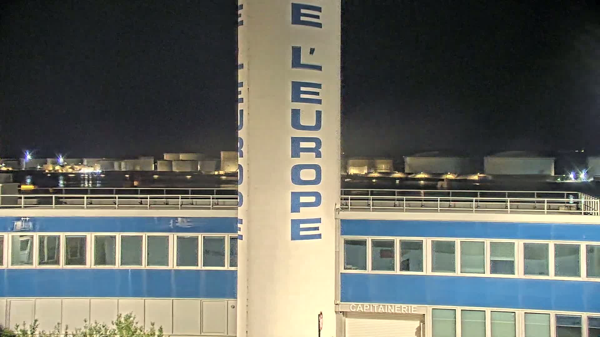 A tall white tower with blue lettering stands in front of a blue building at night, with a body of water and illuminated storage tanks visible in the background.