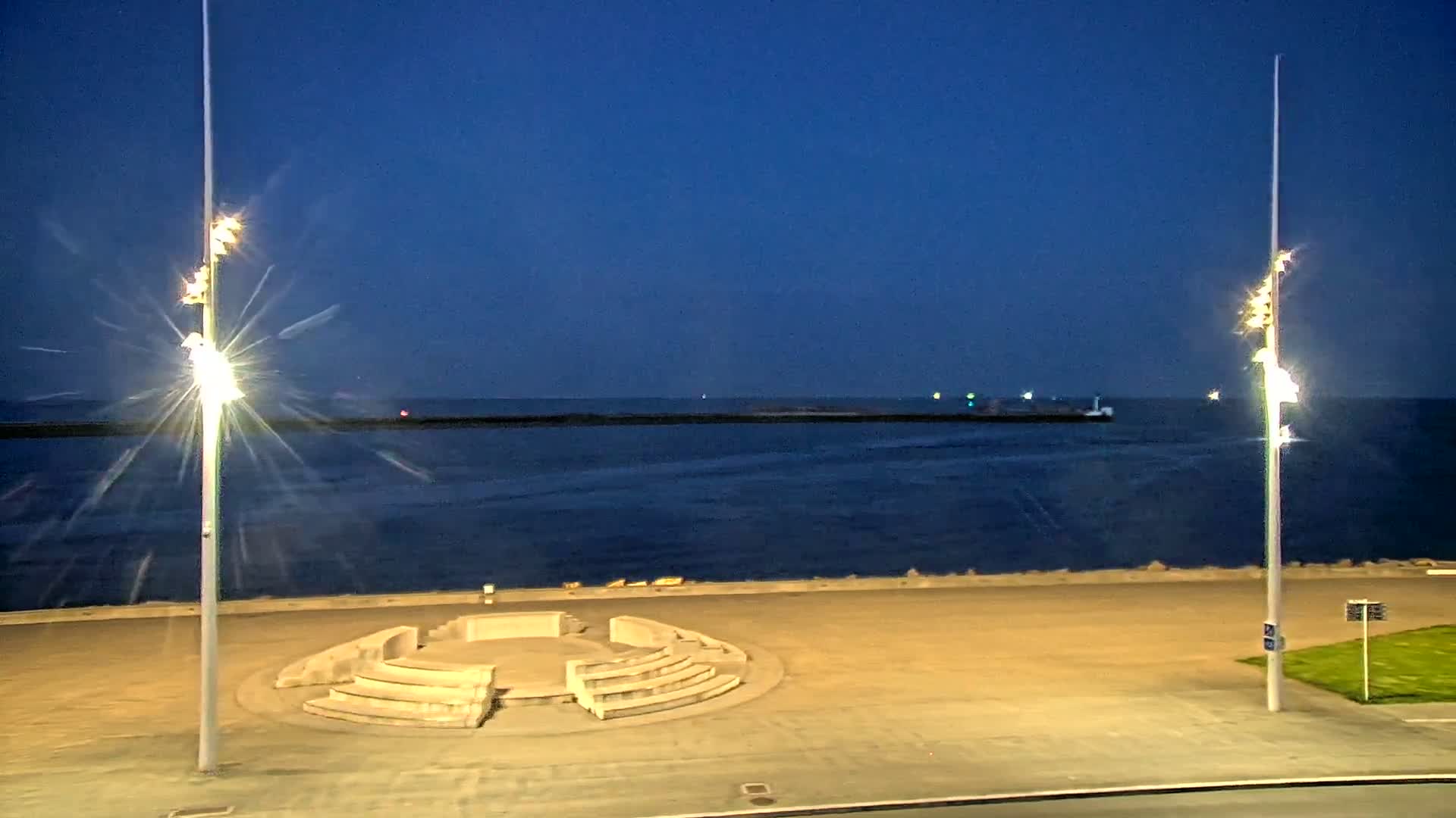Under a dark blue, twilight sky, an empty, circular stone amphitheater sits on a paved waterfront overlooking a calm sea, illuminated by two tall, bright lights.