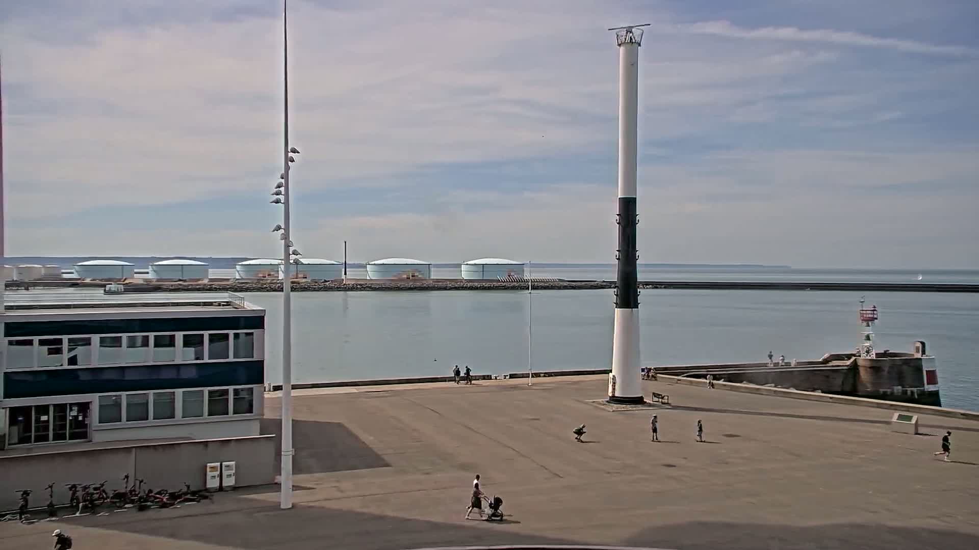 A mostly sunny day shows a harbor with a tall, slender lighthouse, several people walking on a paved area, a building, and several large storage tanks in the background across the water.