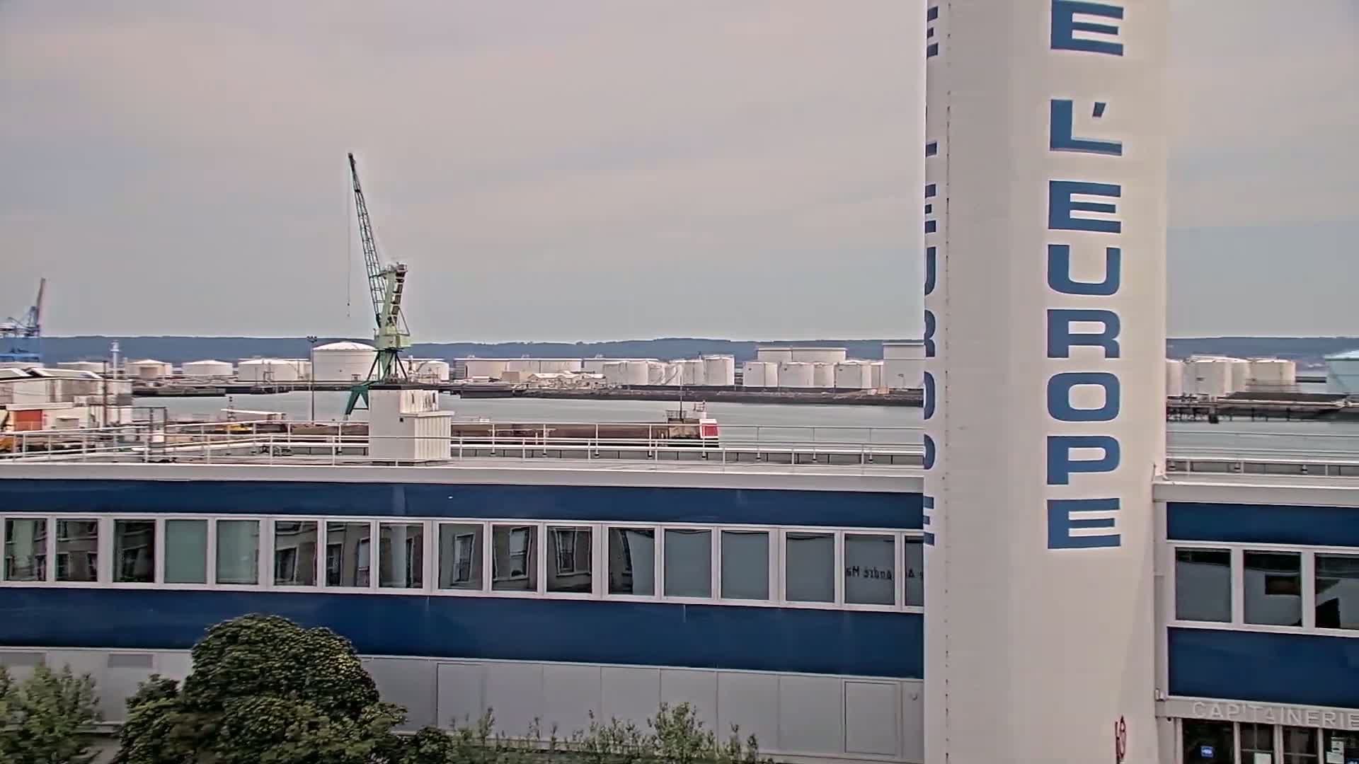 A view of an industrial waterfront on an overcast day, showing a blue building, a crane, and numerous large storage tanks across the water.
