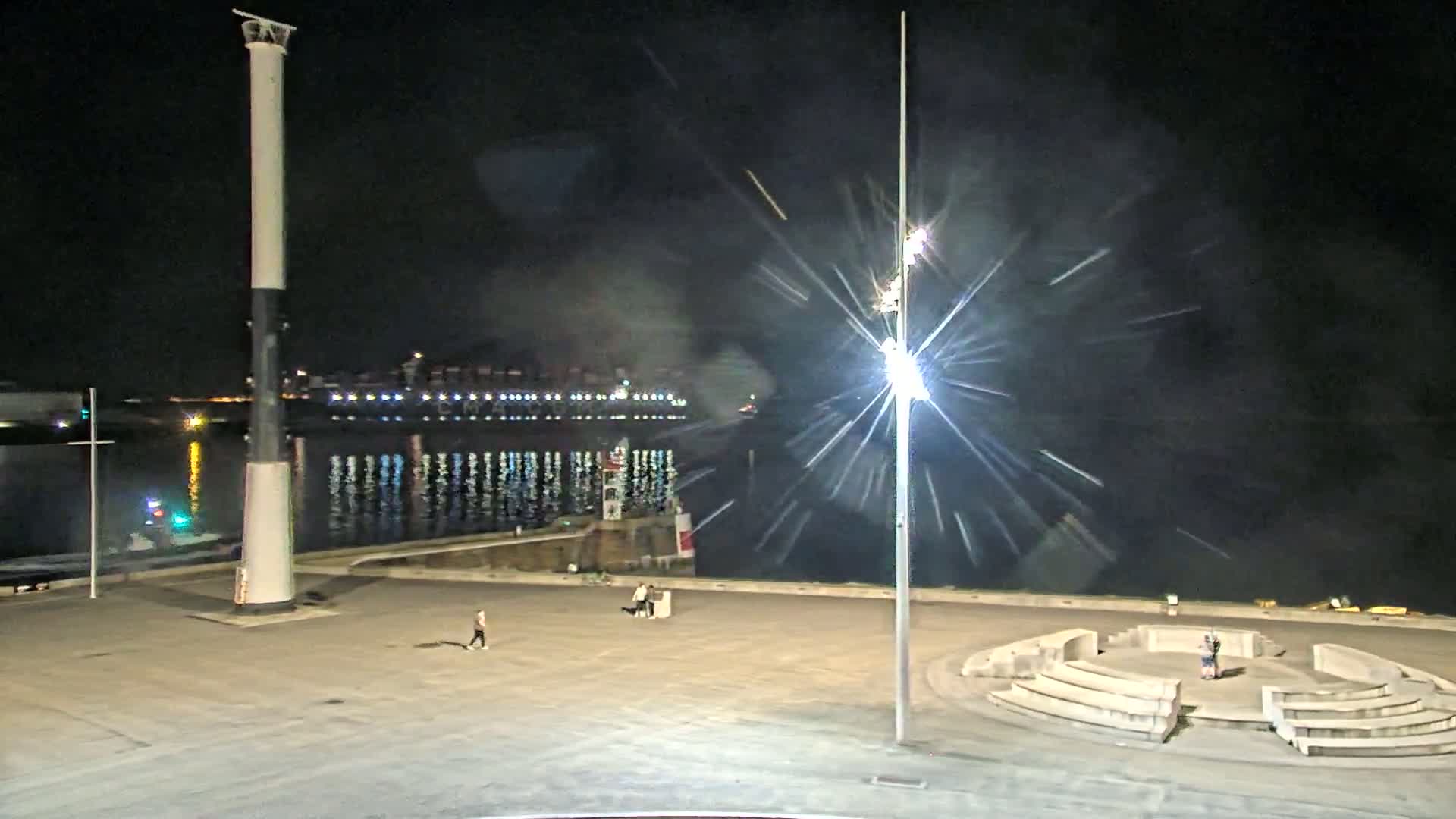 A nighttime view of a harbor with a large ship in the background, a tall column in the foreground, and several people walking in an empty plaza.