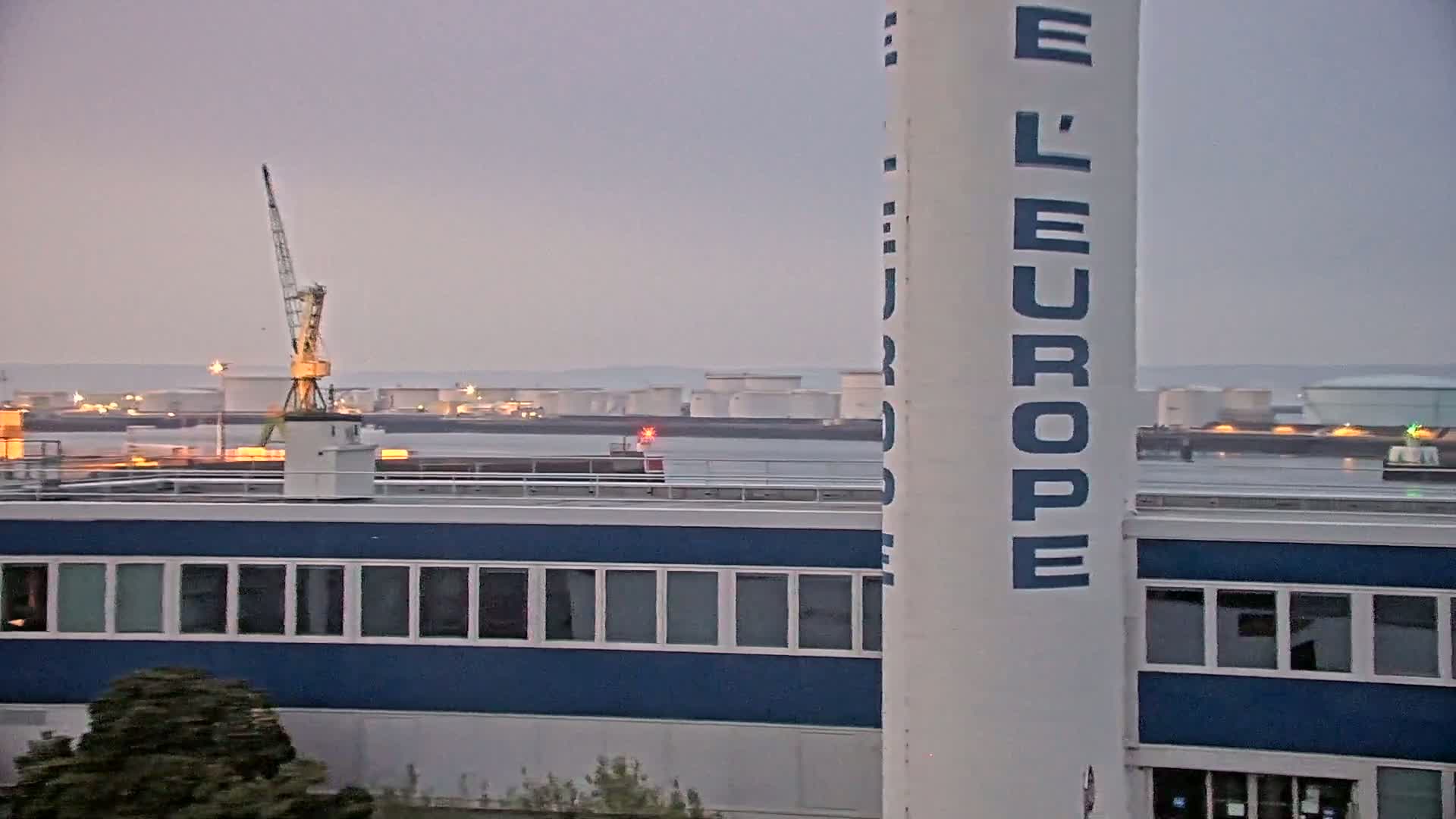 A large, white tower with "L'EUROPE" written vertically on it stands beside a blue building with many windows, overlooking a harbor with a crane and numerous storage tanks under an overcast sky.