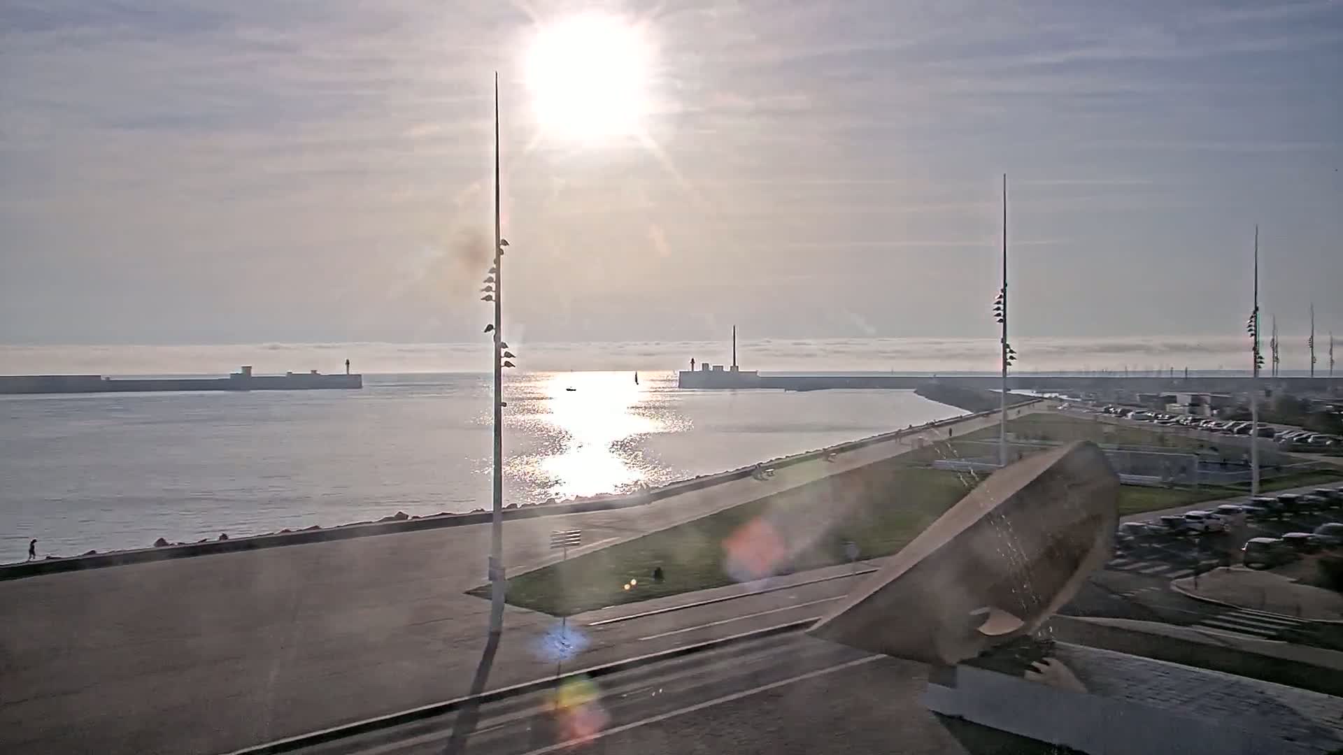 A bright sun shines over a calm harbor, with a long breakwater extending into the distance and a paved waterfront area with a large sculptural element in the foreground.