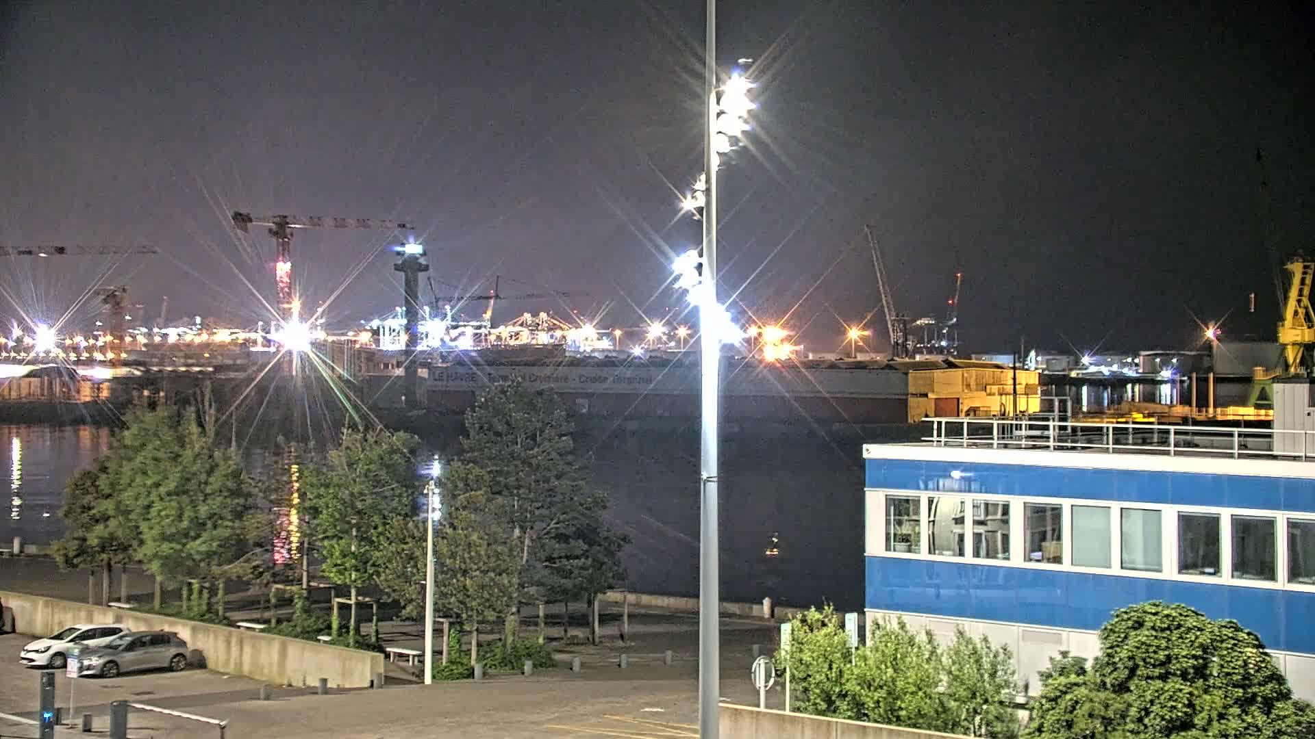A nighttime view of a harbor with brightly lit cargo ships and cranes, a calm body of water reflecting the lights, and a blue building in the foreground.