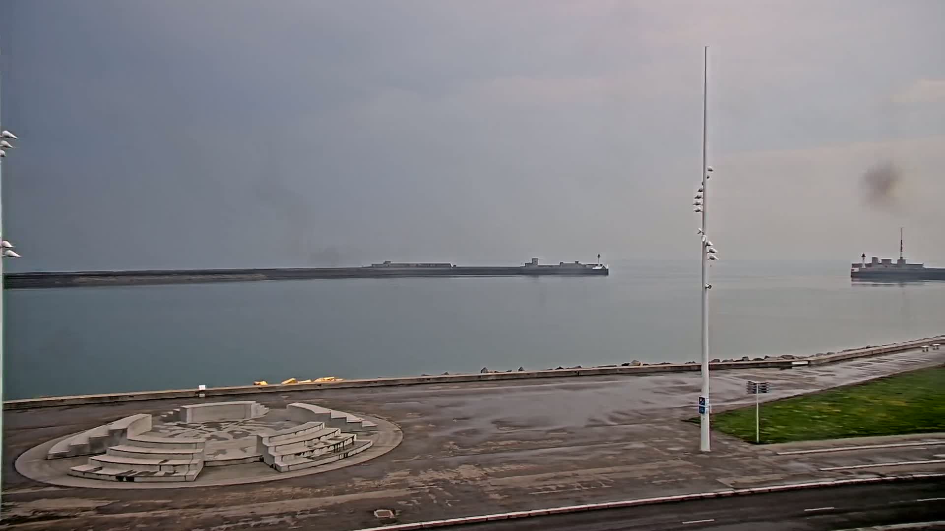 A harbor with calm water under an overcast sky features a circular concrete seating area, a tall pole with lights, and distant breakwaters with small ships.