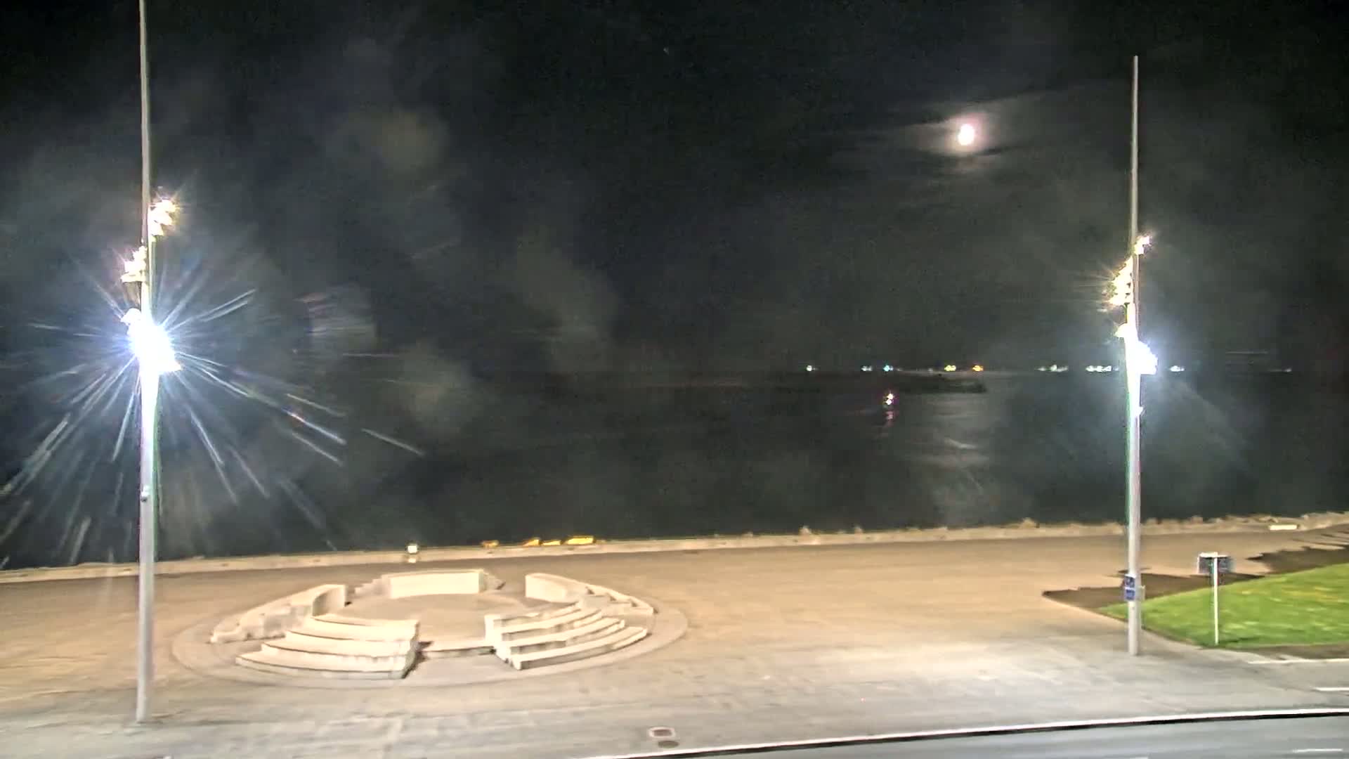 A circular stone seating area sits on a paved waterfront at night under a partly cloudy moonlit sky.