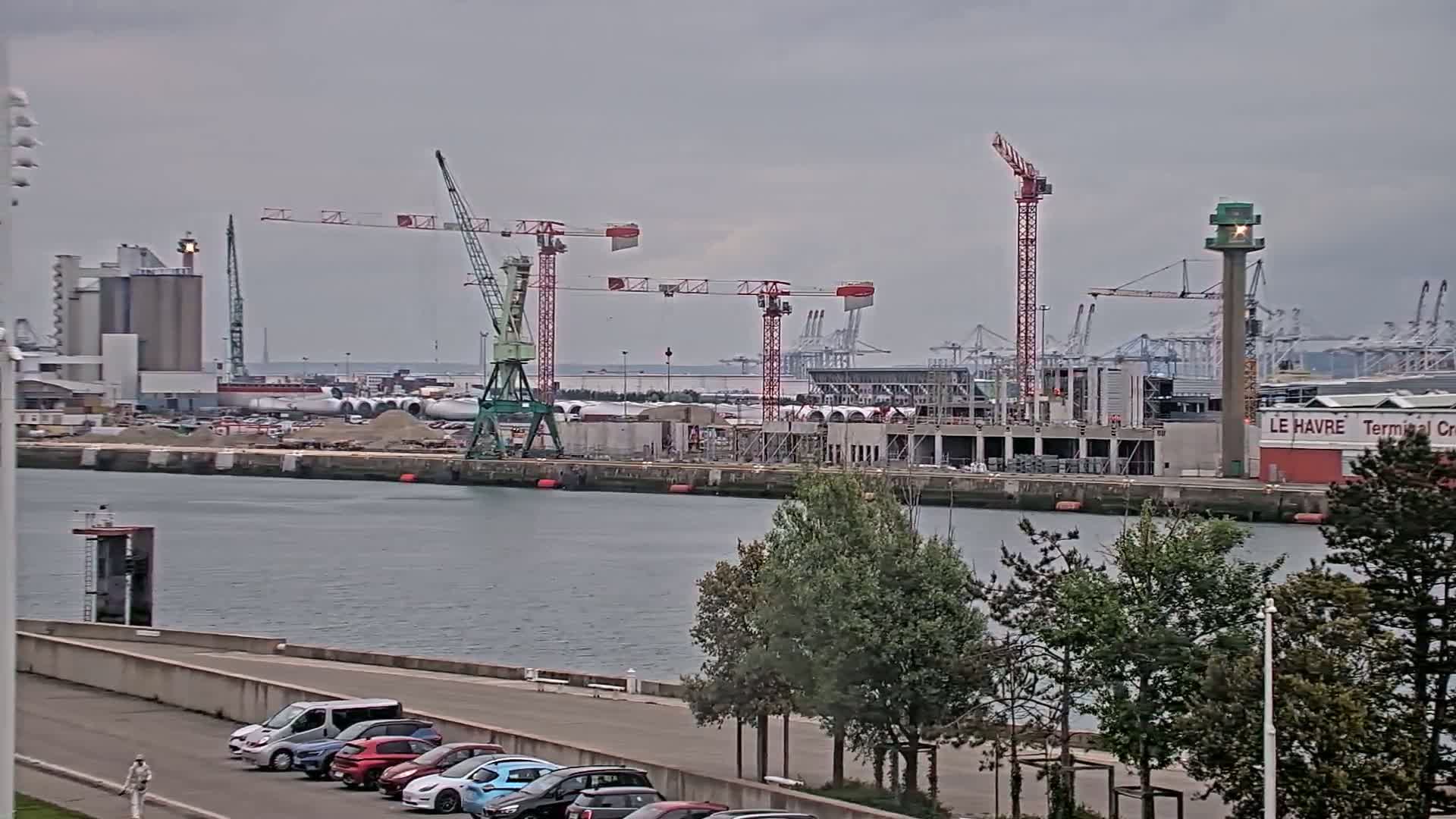 A harbor construction site with several cranes under an overcast sky, and a line of cars parked along a waterfront road in the foreground.