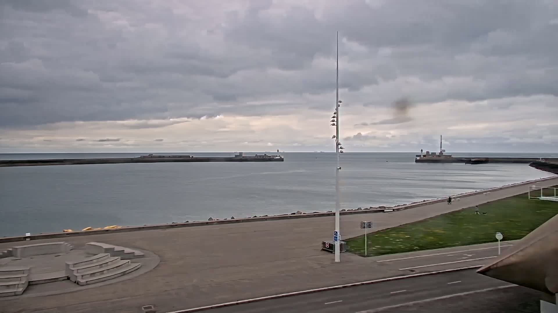 A gray, overcast day shows a harbor with calm water, a paved walkway, and a tall pole with lights at the top.