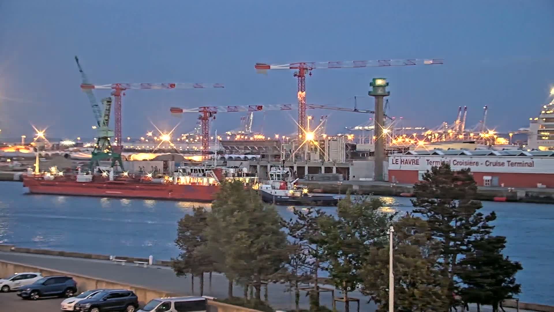 A harbor at dusk, featuring several ships, construction cranes, and a cruise terminal, with lights illuminating the scene.