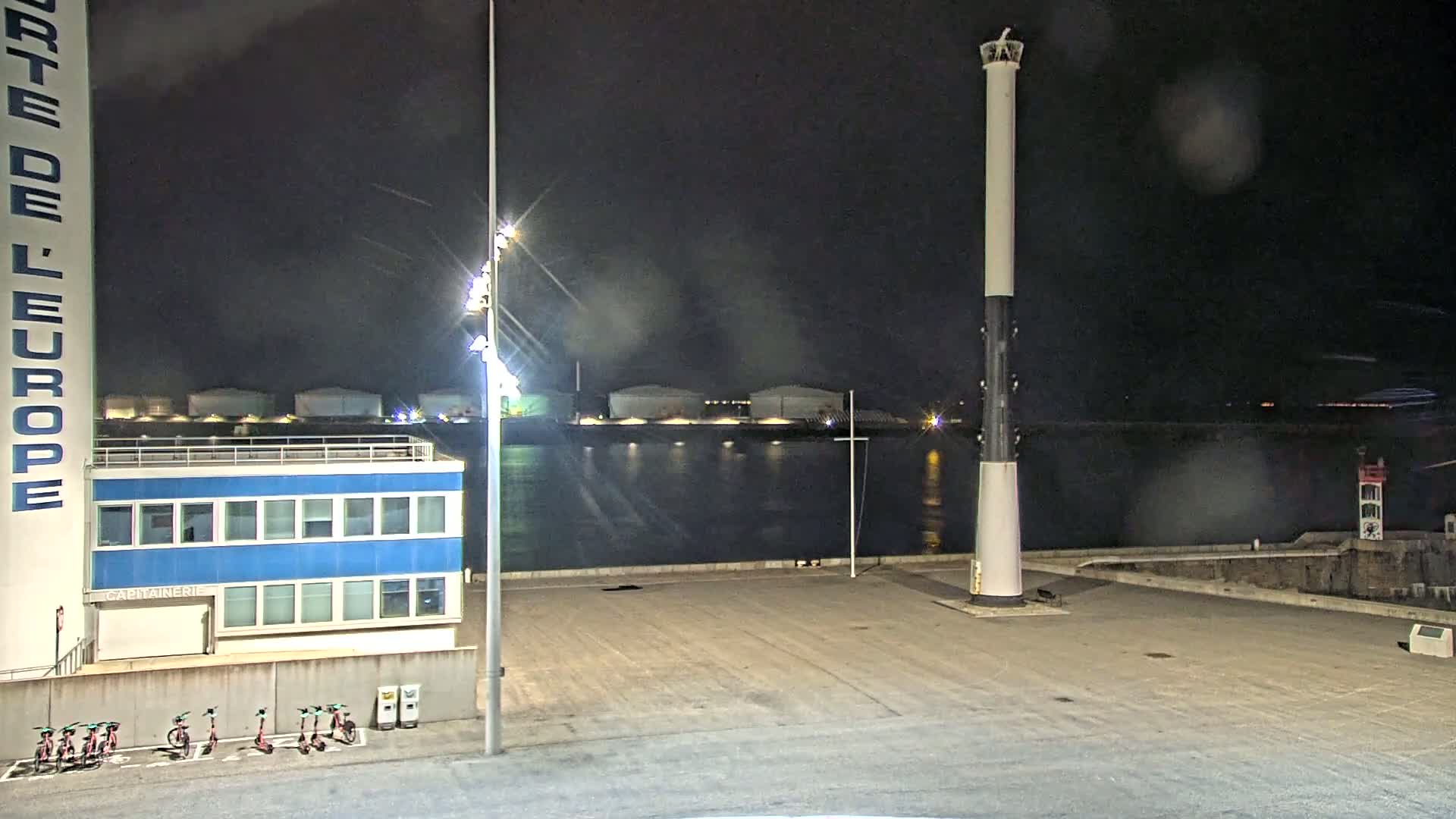 At night, under a dark, slightly rainy sky, a tall lighthouse stands on a dark, empty waterfront area next to a blue building and a row of parked scooters.
