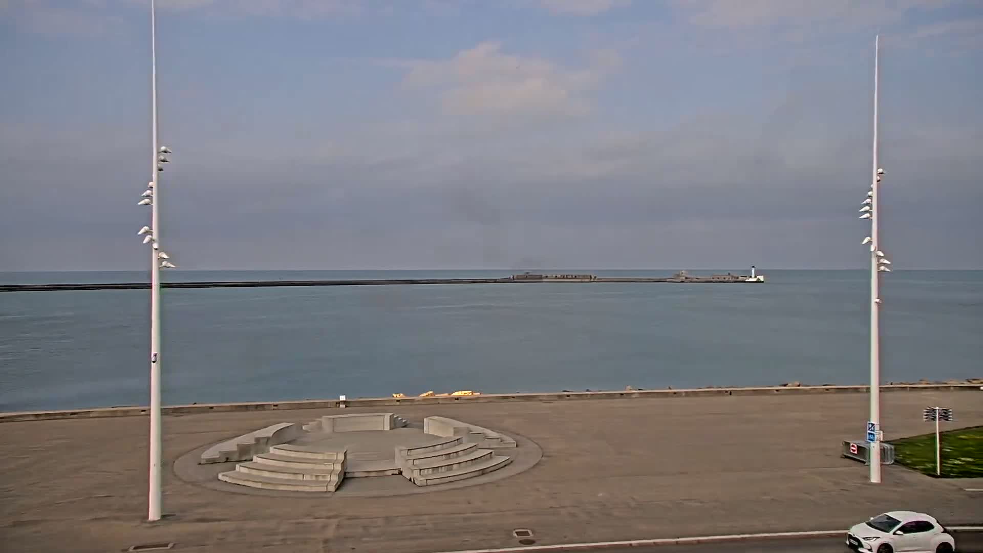 A concrete amphitheater sits on a paved waterfront overlooking a calm sea with a distant breakwater under a mostly cloudy sky.