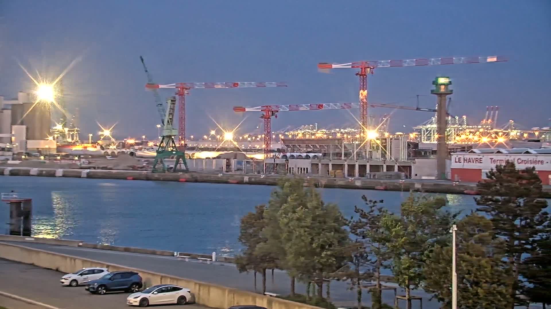 Three large construction cranes stand silhouetted against a twilight sky over a harbor filled with lights and several cars parked along a waterfront path.