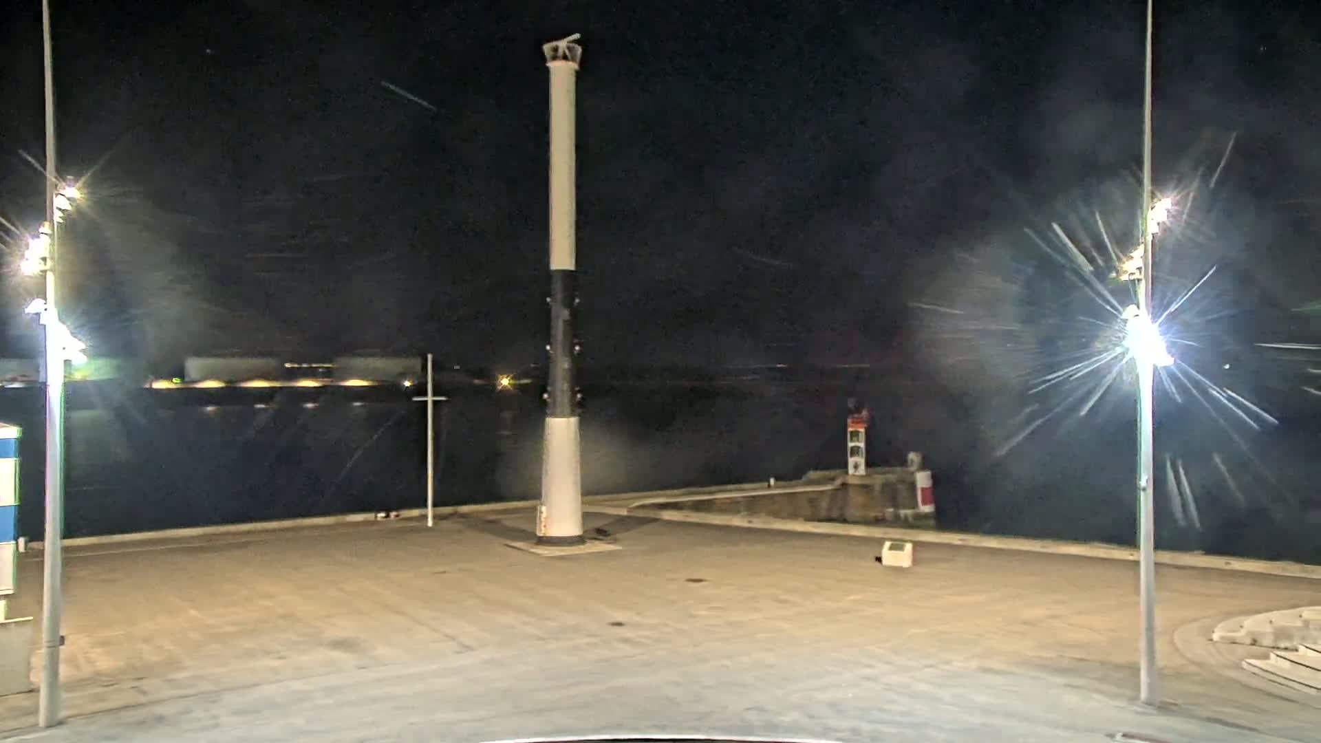 A tall, slender lighthouse stands in a dark, empty paved area at night, with bright lights illuminating the scene and what appears to be snow or heavy rain falling.