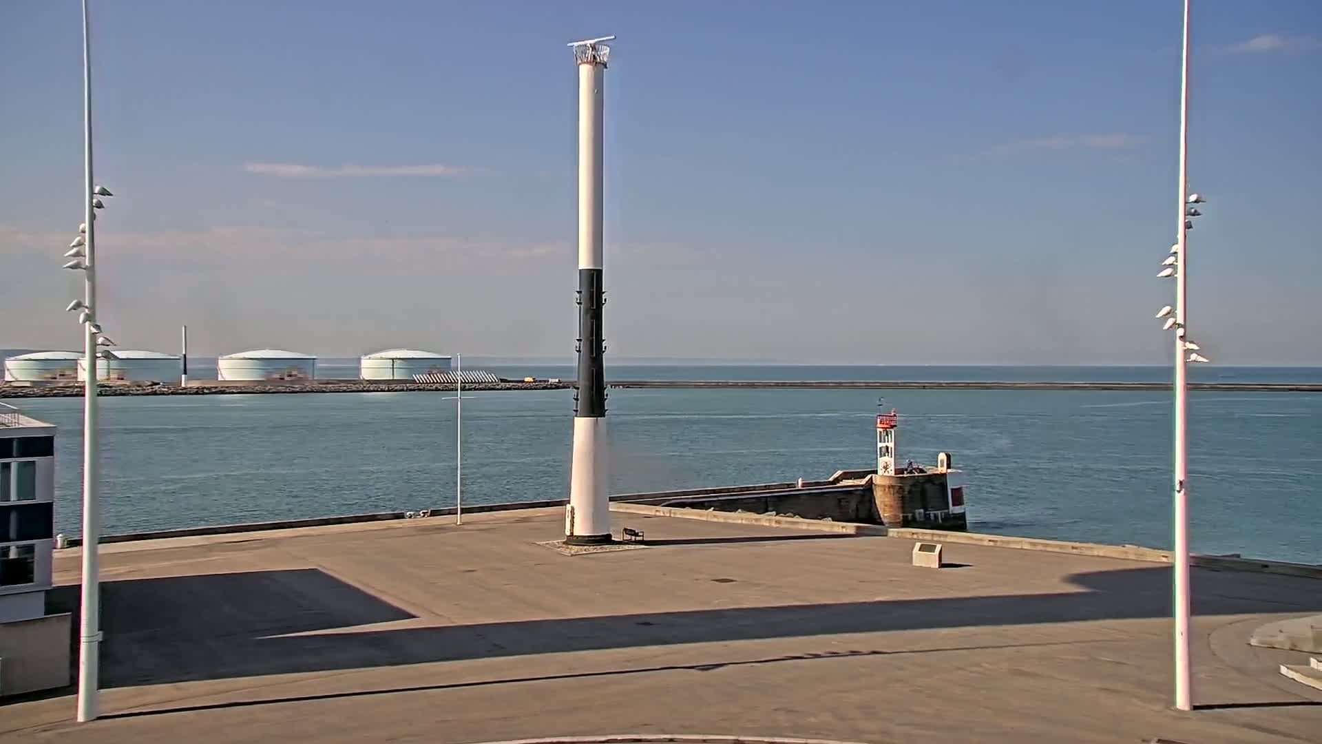 A tall, slender lighthouse with a black band stands in a harbor on a sunny day, with calm water, several storage tanks in the background, and a smaller lighthouse visible in the distance.