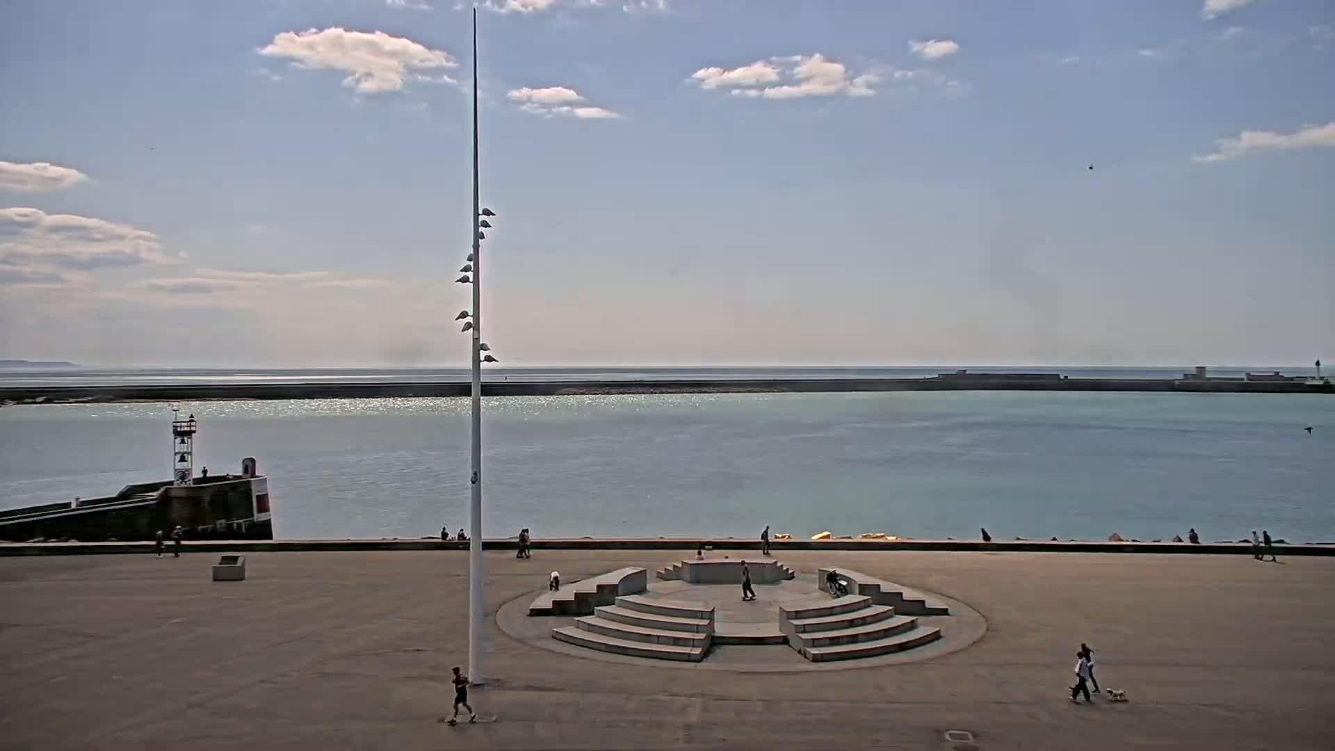 A mostly sunny day shows a harbor scene with a tall flagpole, a small lighthouse, a stone amphitheater-like structure, and a few people walking dogs and skateboarding in a plaza overlooking calm water.