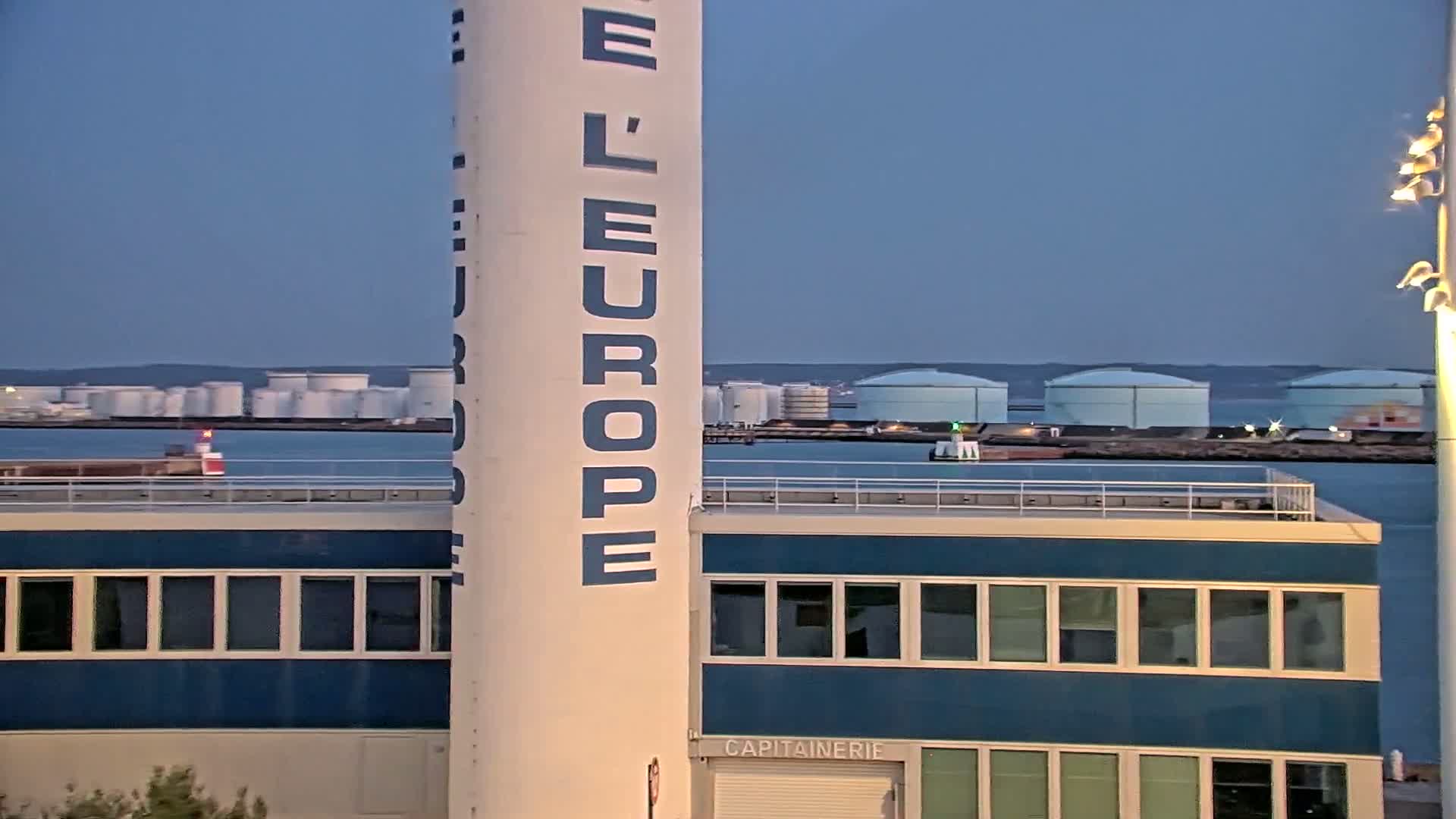 A tall white tower with "L'EUROPE" written vertically on it stands next to a blue building overlooking a calm body of water with several large white storage tanks in the distance under a twilight sky.