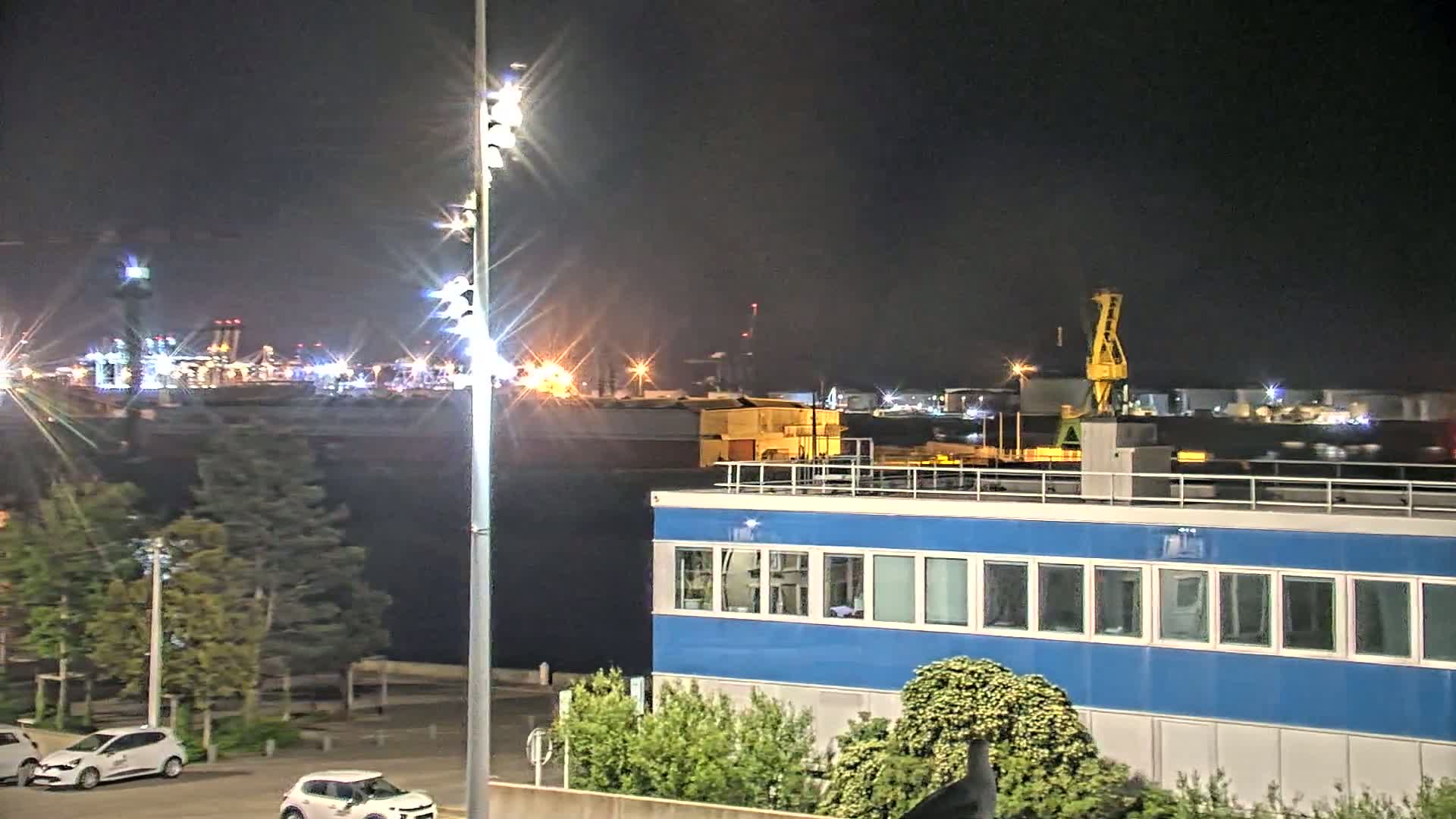 A nighttime view of a harbor with a blue building in the foreground, several cars parked nearby, and industrial structures and a crane visible across the water under a dark sky.