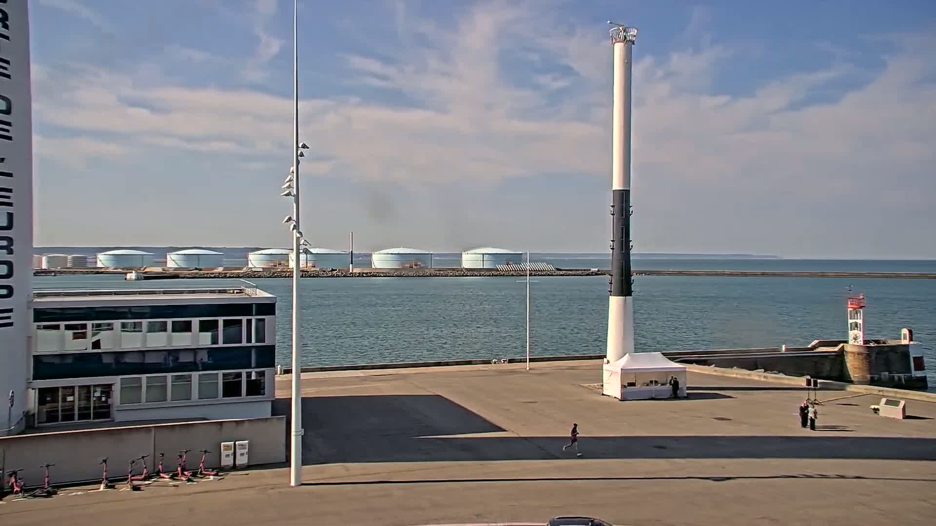 A harbor scene on a partly sunny day shows a body of water with several large storage tanks in the distance, a tall pole, a small building, and a few people.