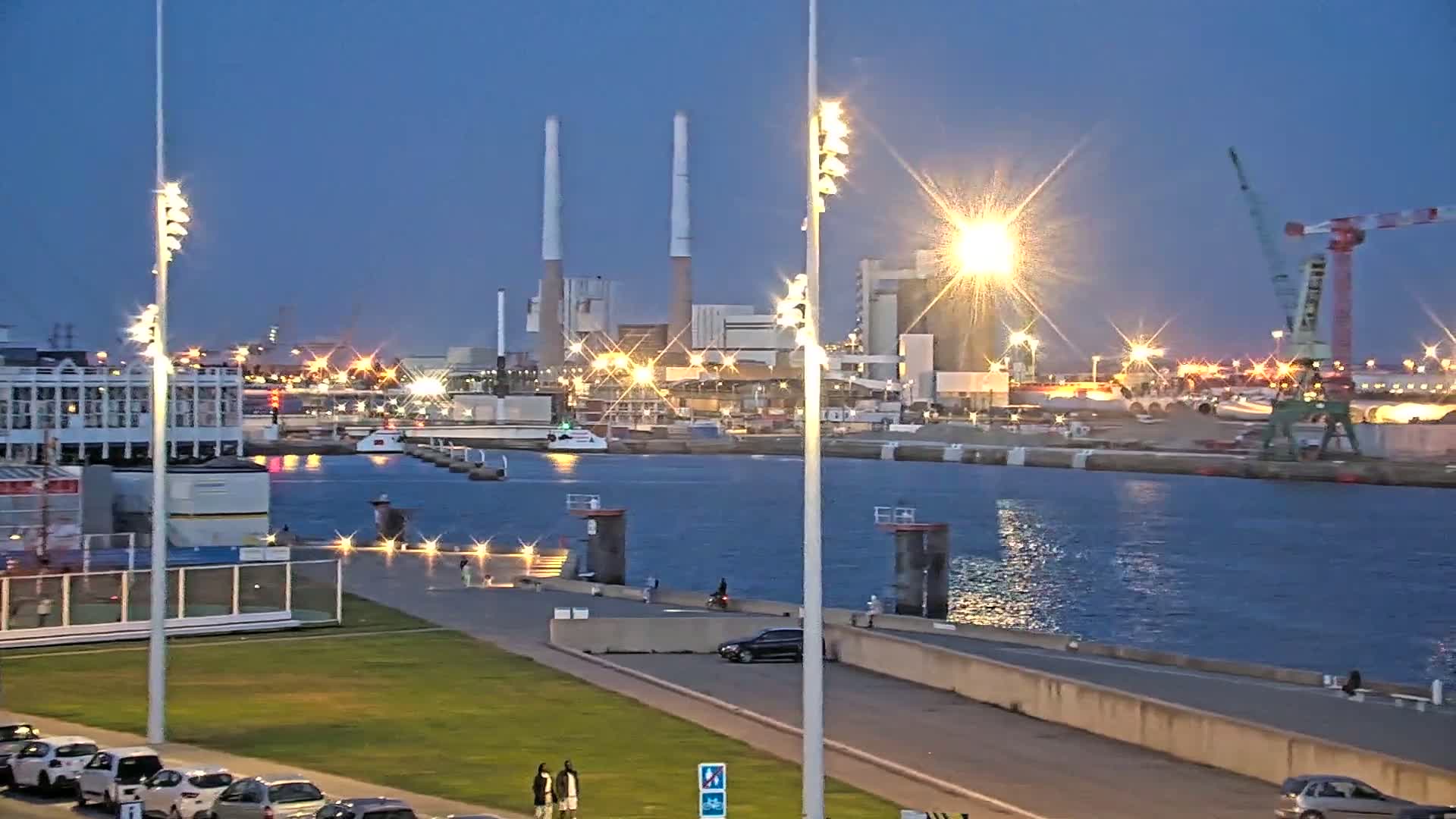 A nighttime view of a harbor with a factory in the background, illuminated by bright lights, and a calm body of water reflecting the lights.