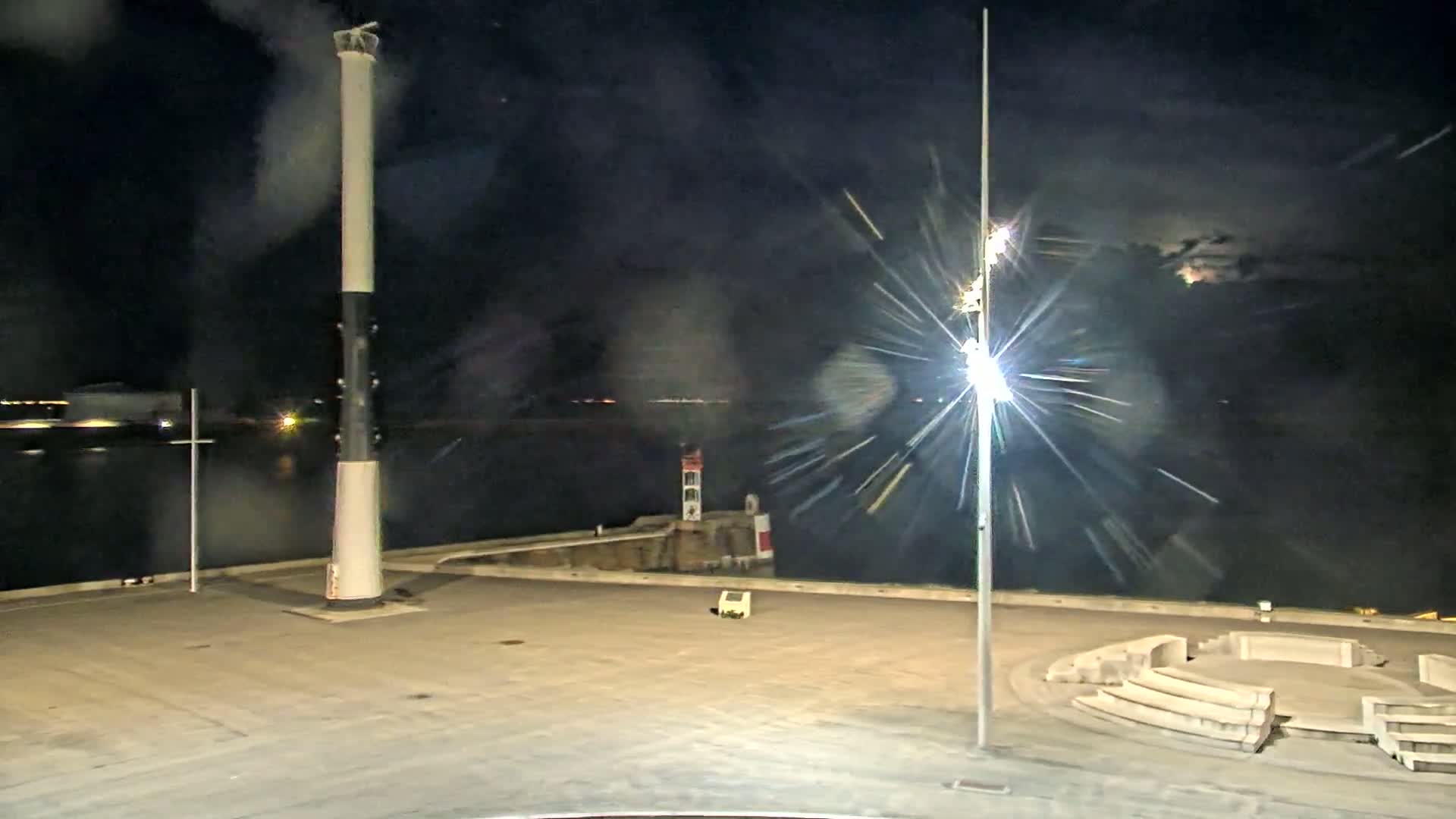 A nighttime view of a harbor shows a tall lighthouse, a smaller lighthouse in the distance, and a circular stone seating area, all under a partly cloudy sky.
