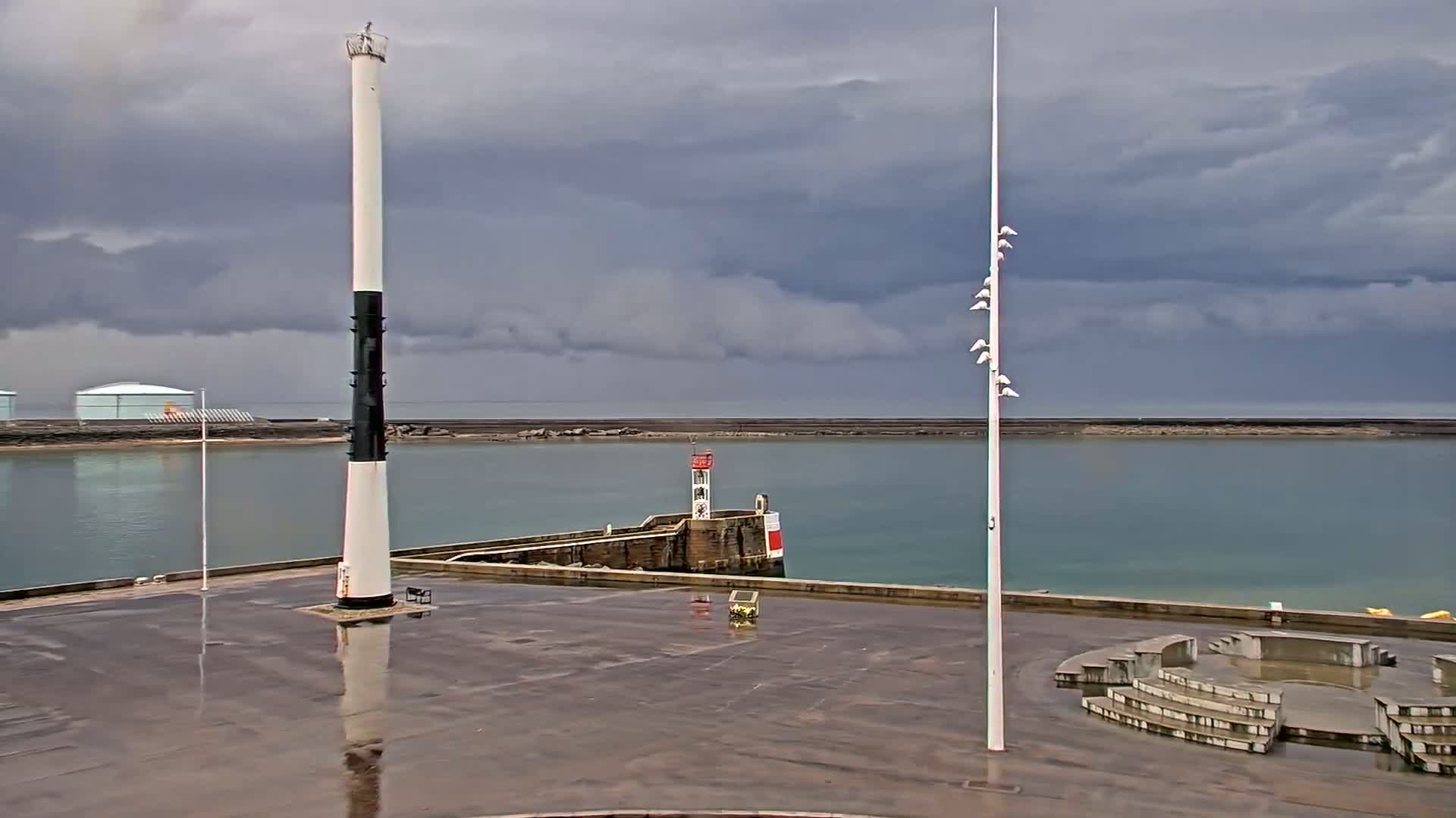 A wet harbor plaza features a tall black and white lighthouse, a smaller lighthouse on a breakwater, and a tall light post under a cloudy sky.