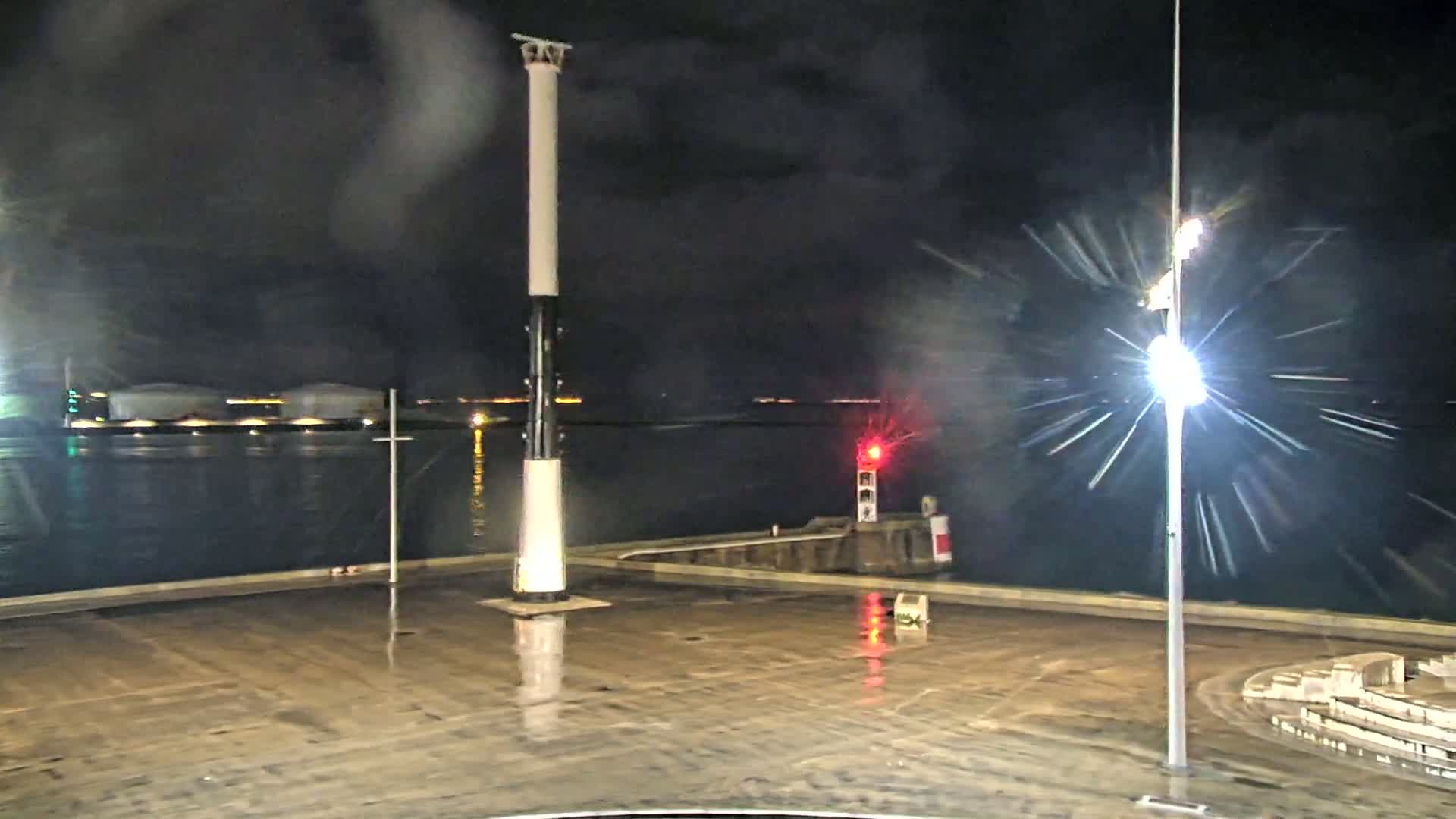 A nighttime view of a wet harbor with a tall pole, a small lighthouse, and large storage tanks in the background under rainy conditions.